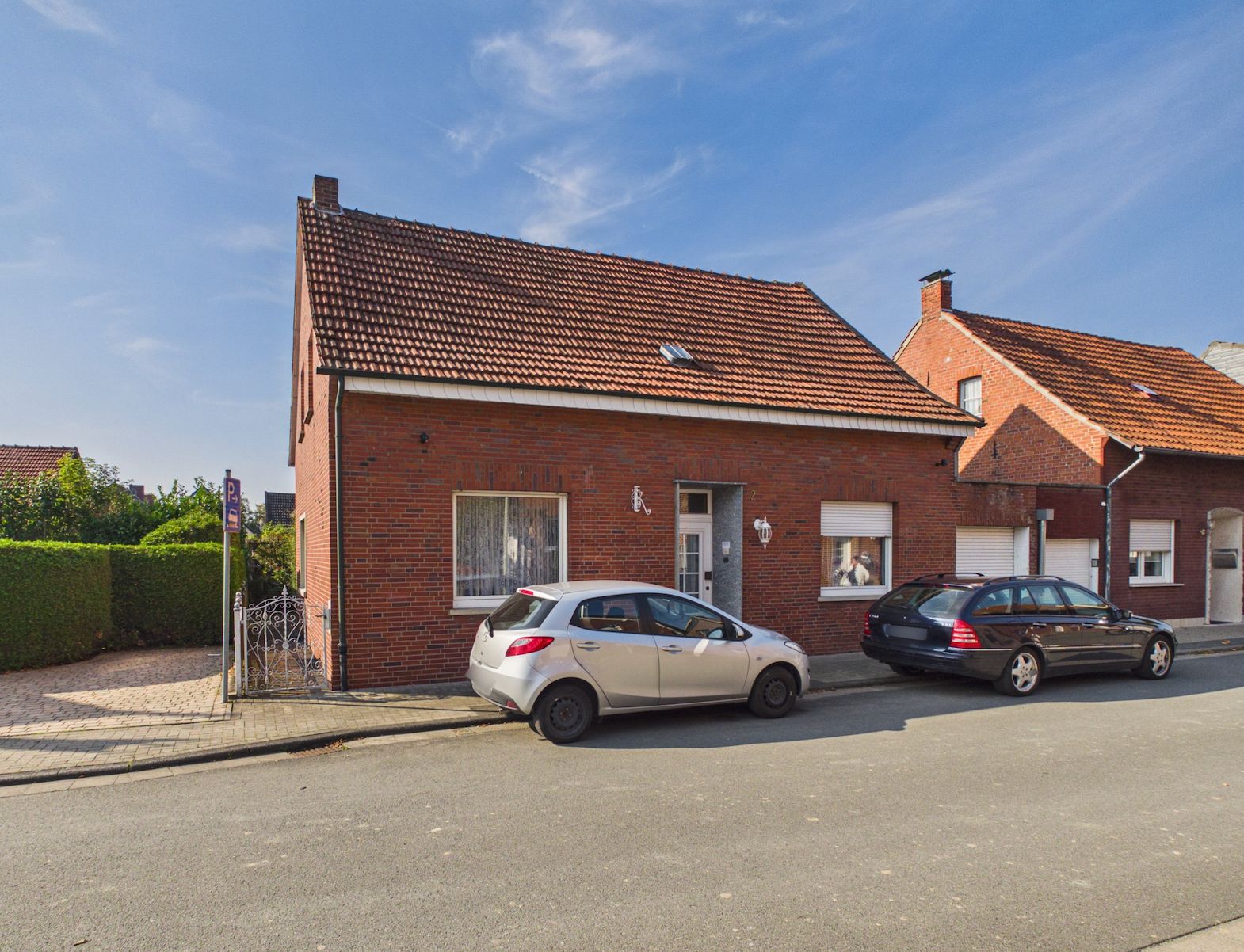 Backsteinwohnhaus mit rotem Dach und zwei geparkten Autos vor dem Haus an einer Straße.