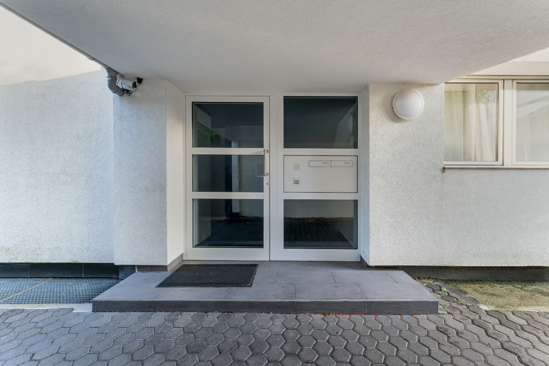 Eingangsbereich eines Wohnhauses mit Glastür, Briefkästen und Fußmatte.