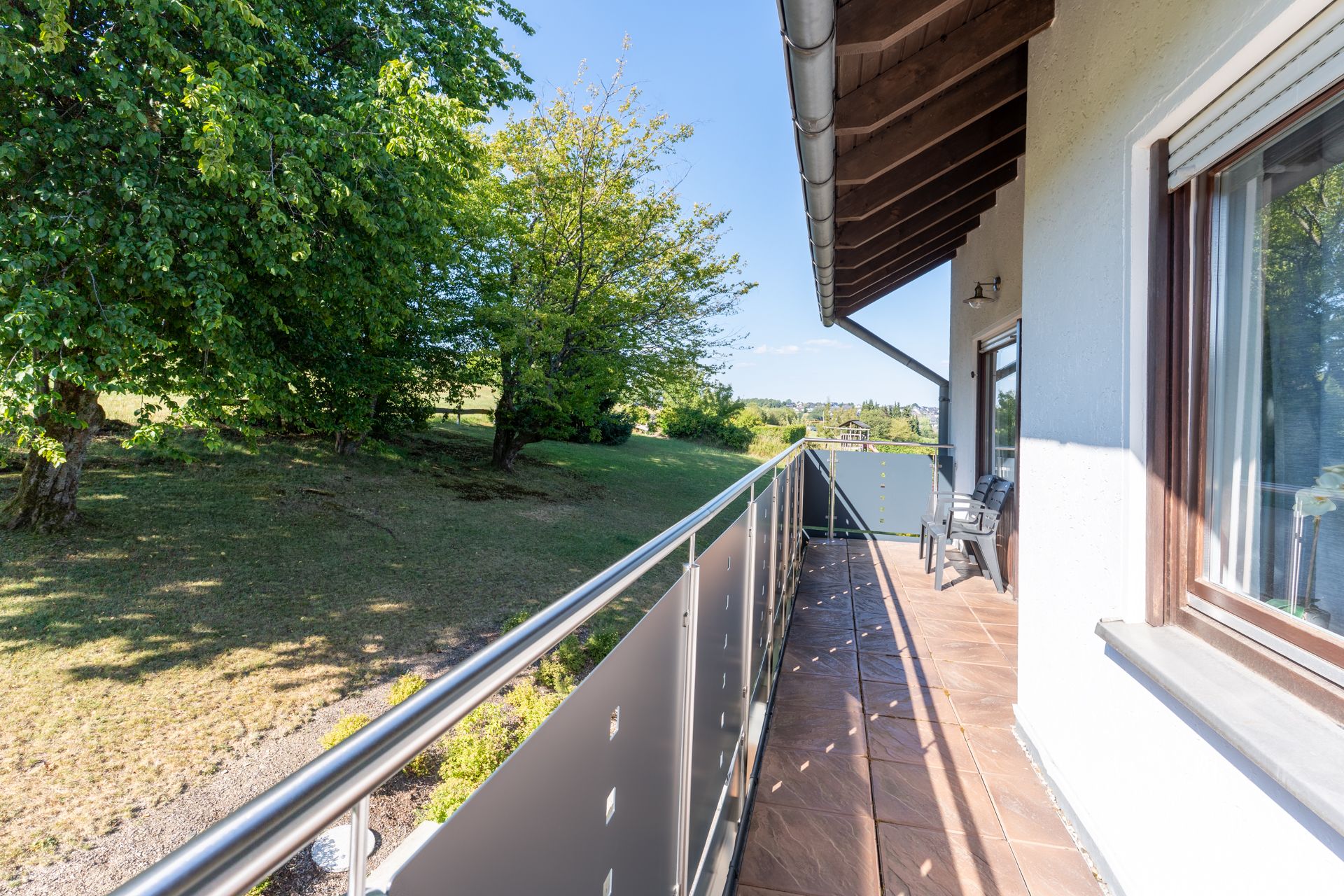 Langer Balkon mit Glasgeländer und Blick auf einen Garten mit Bäumen.