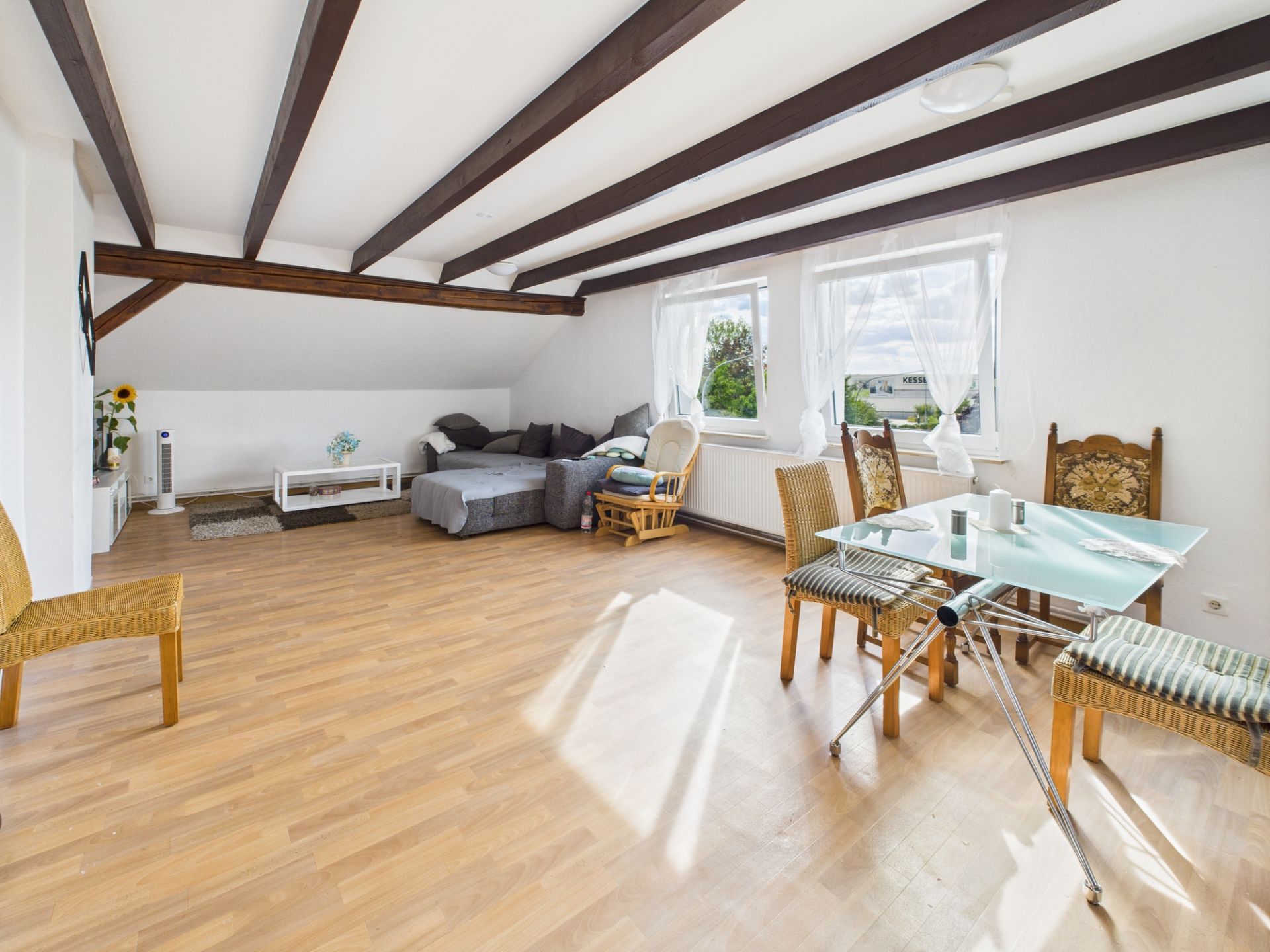 Helles Wohnzimmer mit Holzboden, Glas-Esstisch, Stühlen und Sofa unter Dachbalken.
