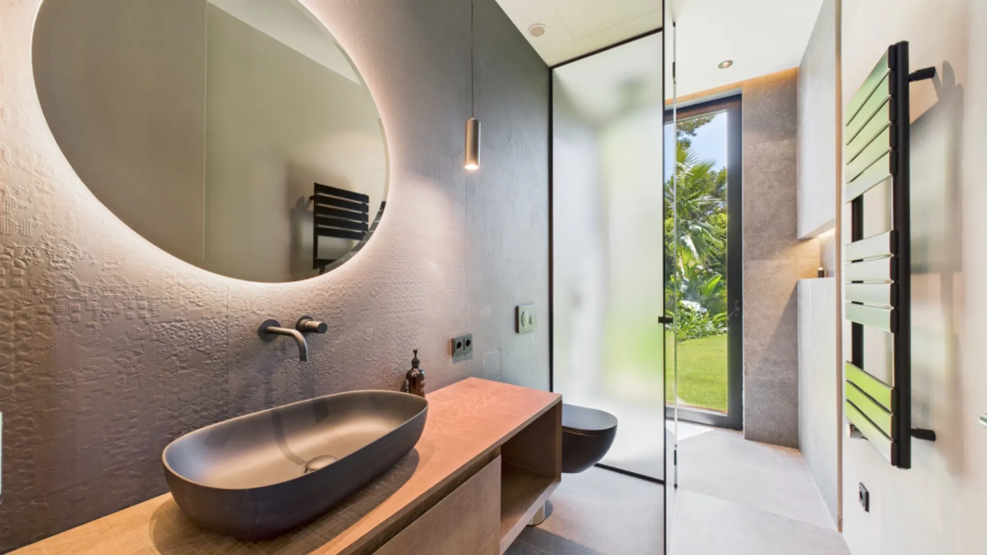 Modernes Badezimmer mit rundem Spiegel, Waschbecken und großer Glastür zum Garten.
