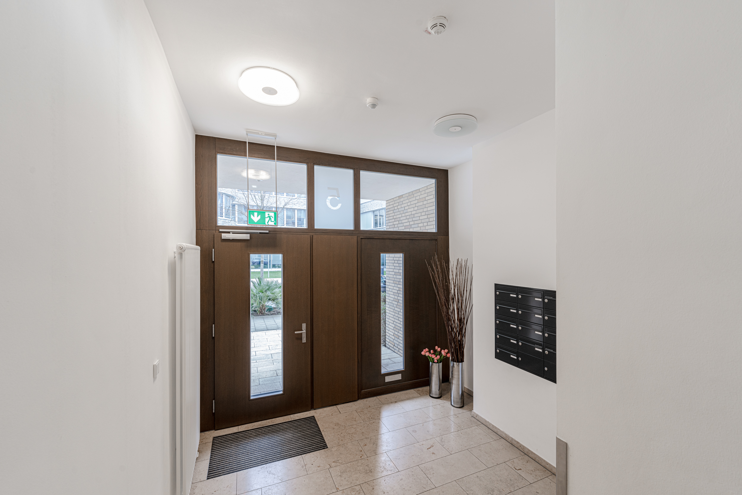Heller Hausflur mit Holztür, Briefkästen, Pflanzen und Fliesenboden.