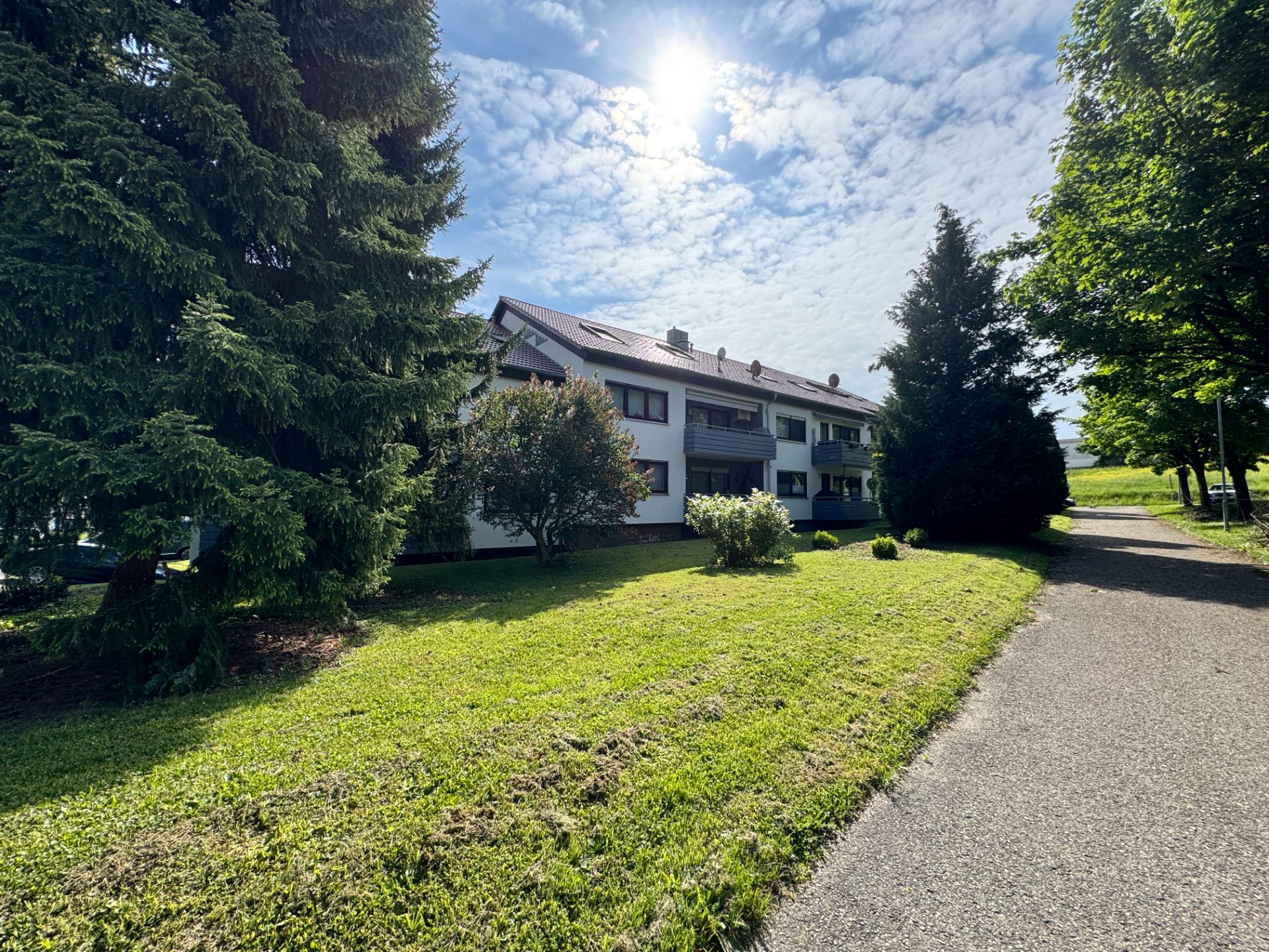 Wohnhaus mit Balkon und Garten, umgeben von Bäumen, bei sonnigem Wetter.