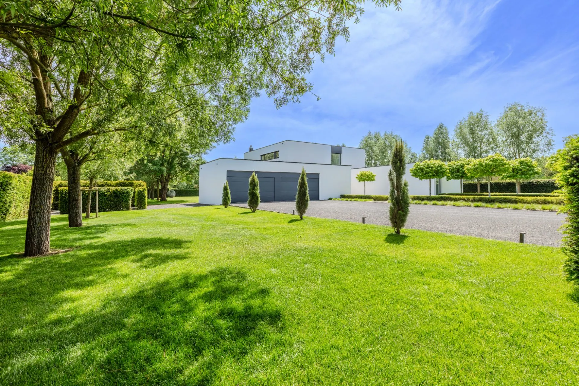 Modernes weißes Wohnhaus mit großem Garten und Einfahrt, umgeben von Bäumen.