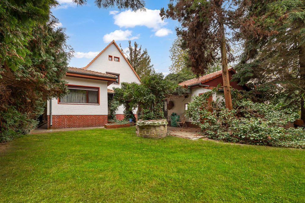 Haus mit rotem Dach und großem Garten, umgeben von Bäumen und Sträuchern, mit einem alten Brunnen in der Mitte.