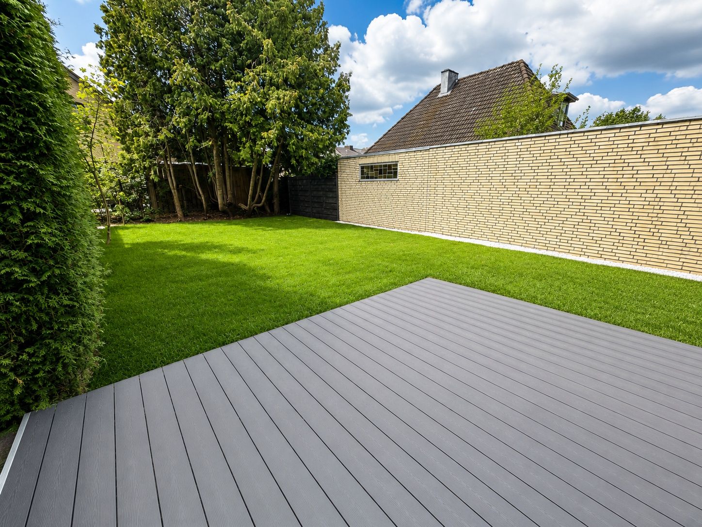 Garten mit grauer Terrasse, grünem Rasen, Hecke und Bäumen, begrenzt von einer Mauer.