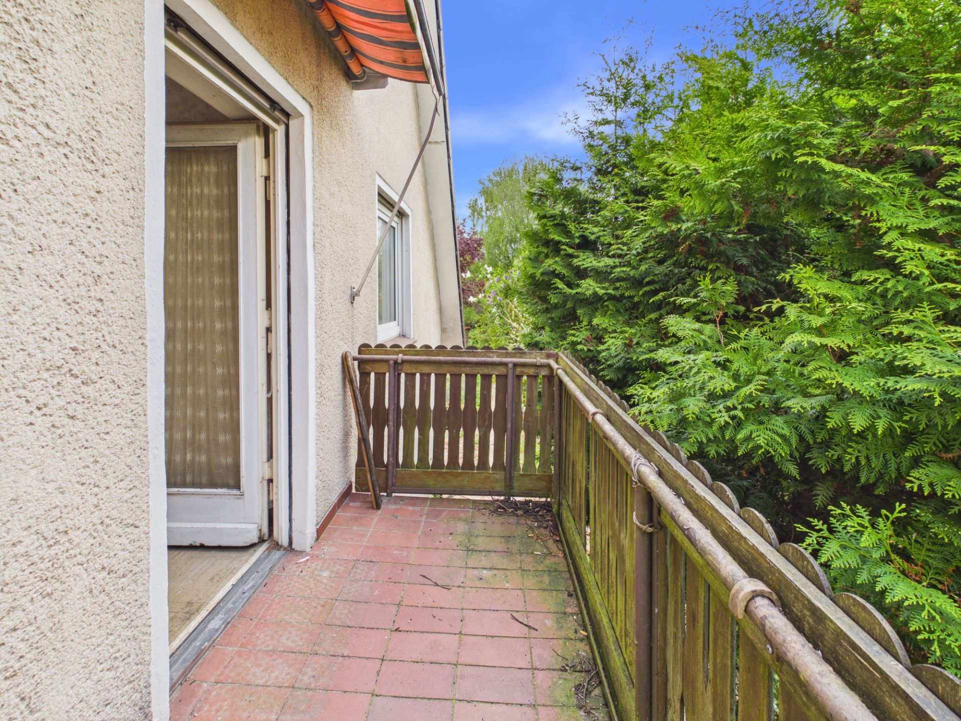 Kleiner Balkon mit Holzgitter und Markise, daneben dichte grüne Bäume.