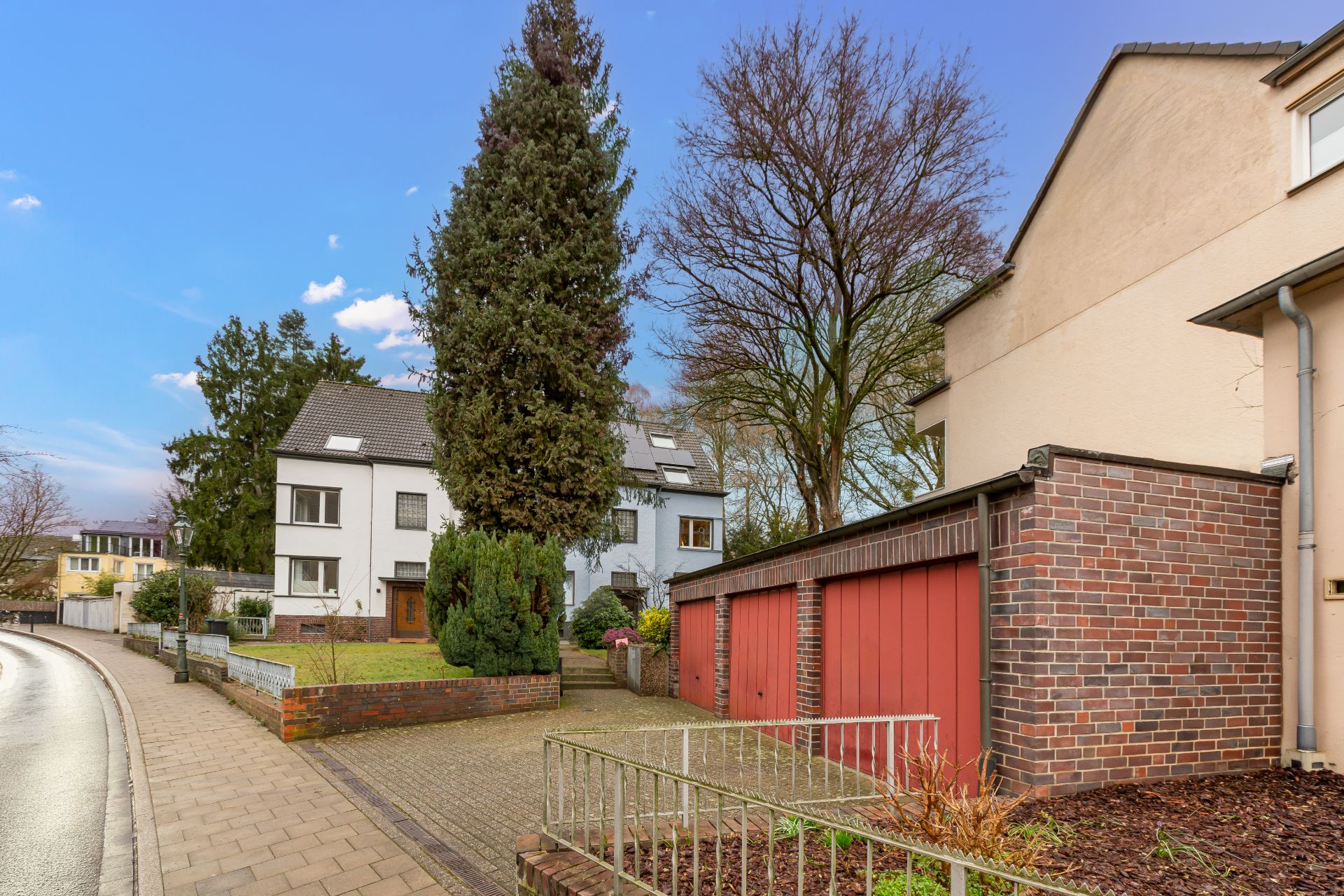 Wohnstraße mit Mehrfamilienhaus, roten Garagen und Bäumen bei klarem Himmel.