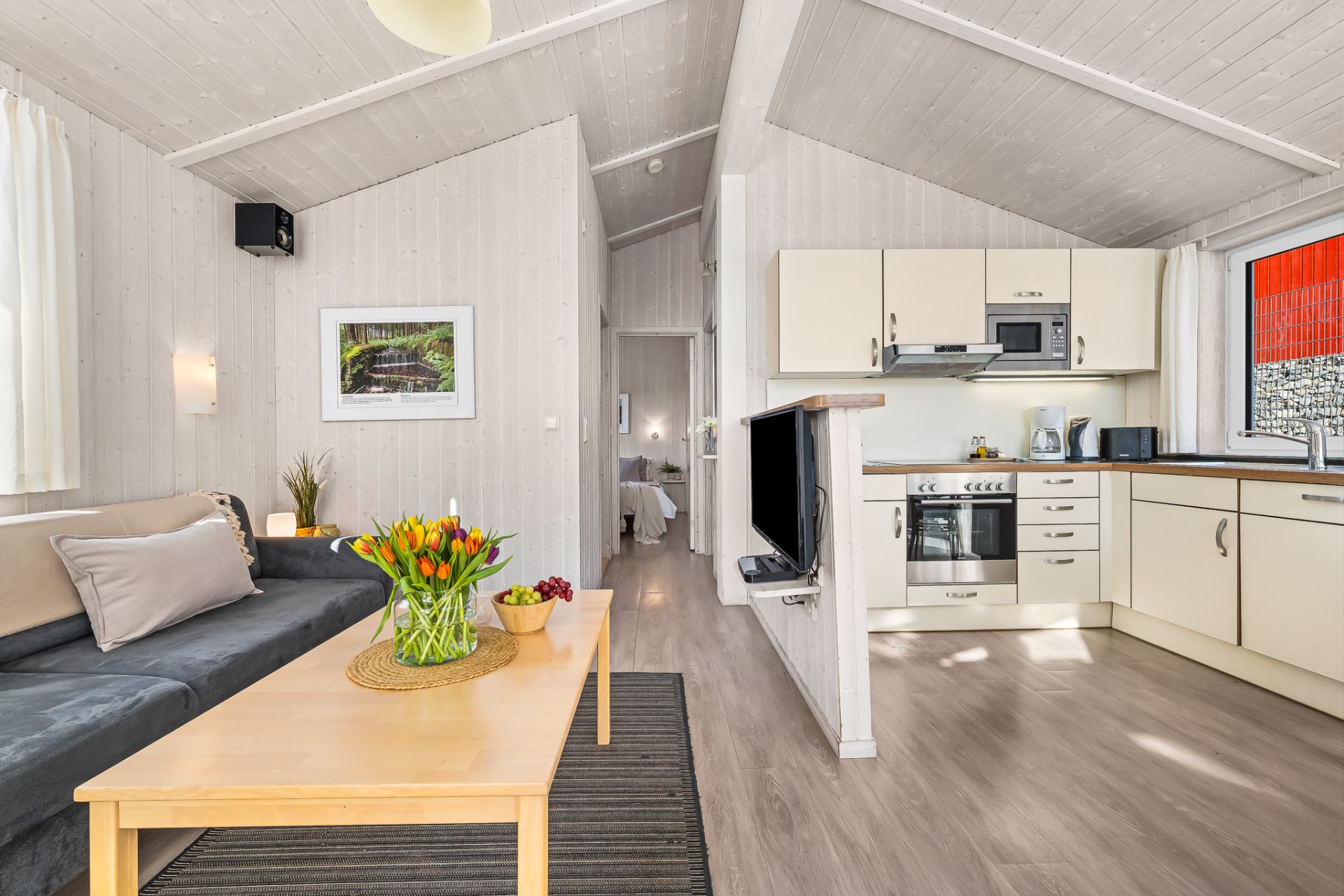 Helles Wohnzimmer mit Sofa, Couchtisch, Blumen und offener Küche in einem Haus.
