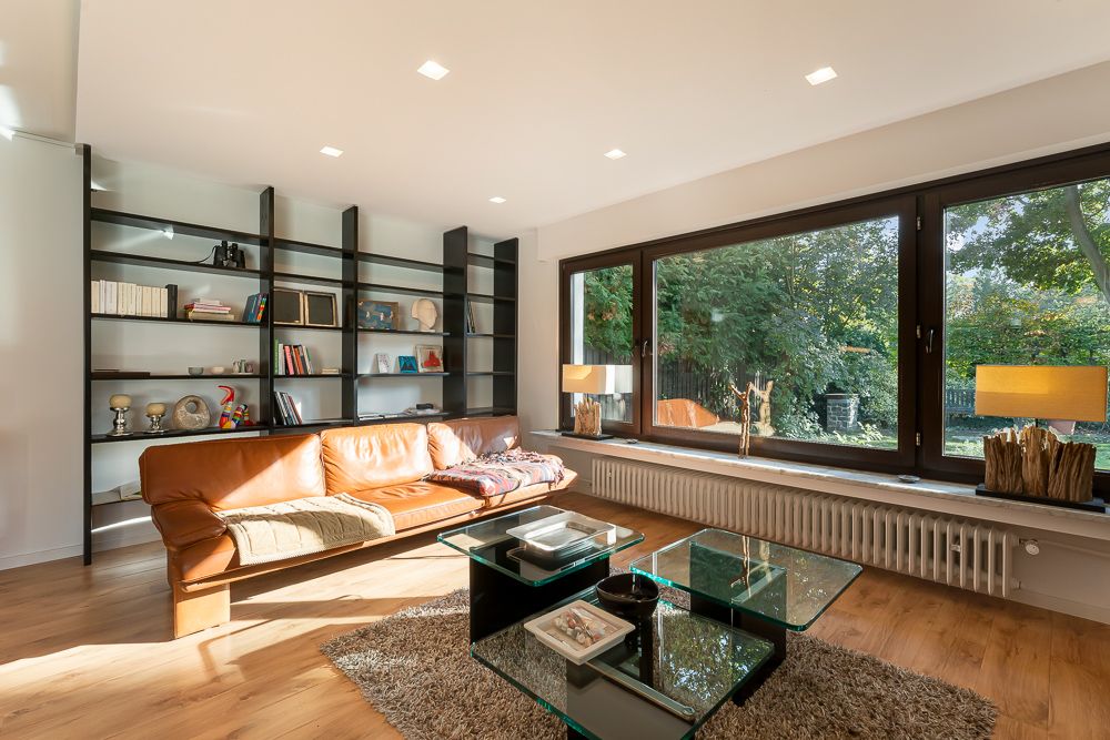 Helles Wohnzimmer mit braunem Sofa, Regalwand, Glastisch und großem Fenster mit Blick ins Grüne.