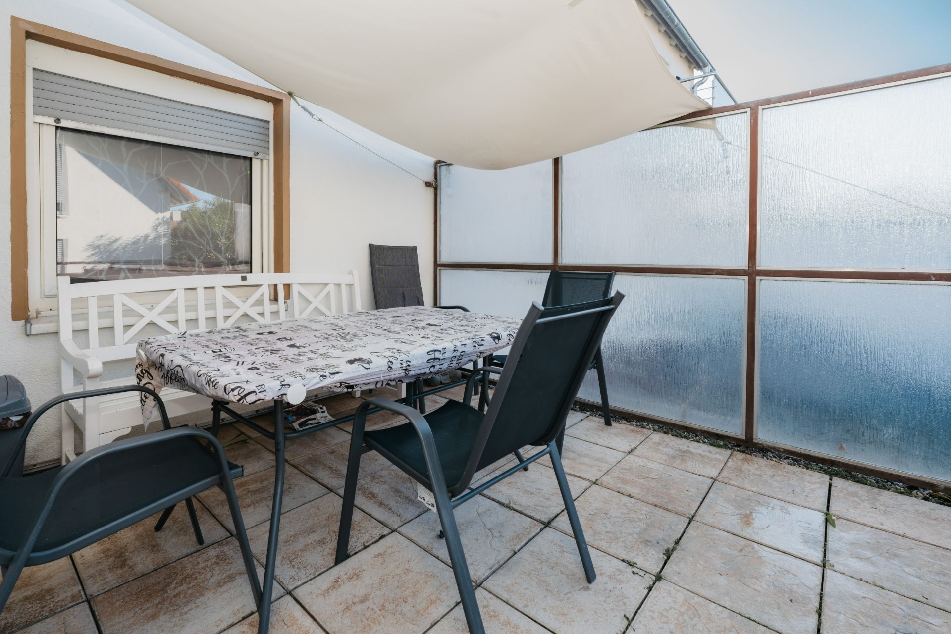 Überdachte Terrasse mit Tisch, Stühlen und Bank auf gefliestem Boden, Sichtschutzwände vorhanden.