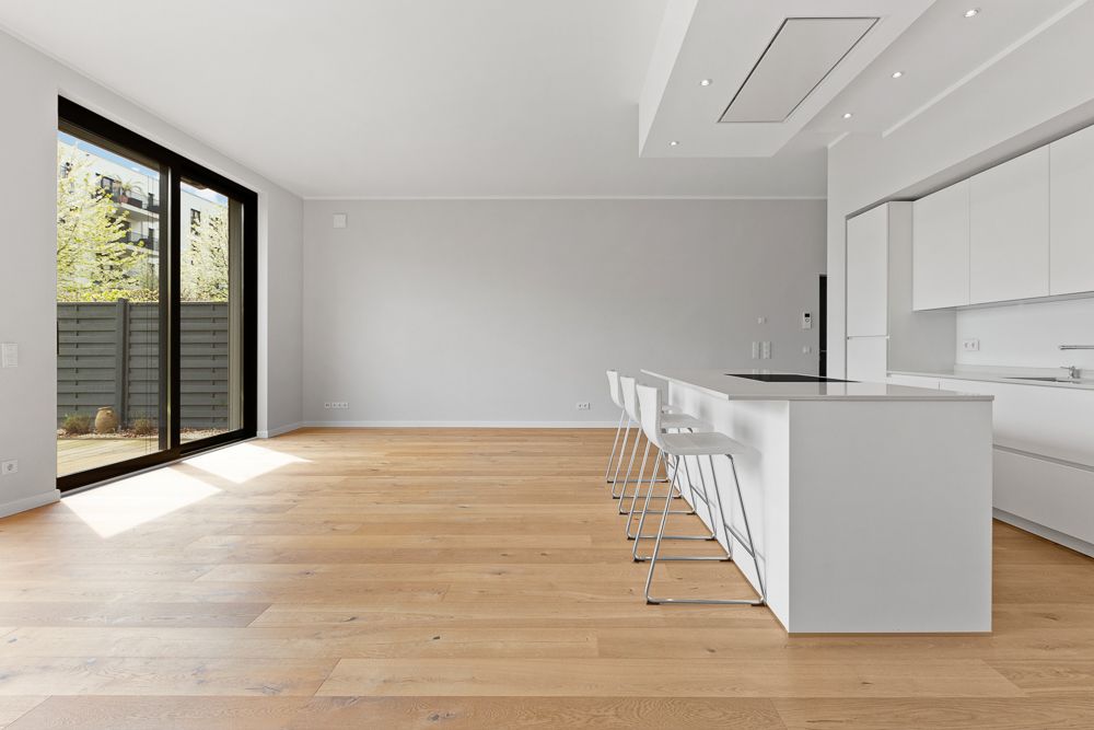 Heller Raum mit Holzfußboden, großer Fensterfront und moderner weißer Küche mit Kücheninsel.