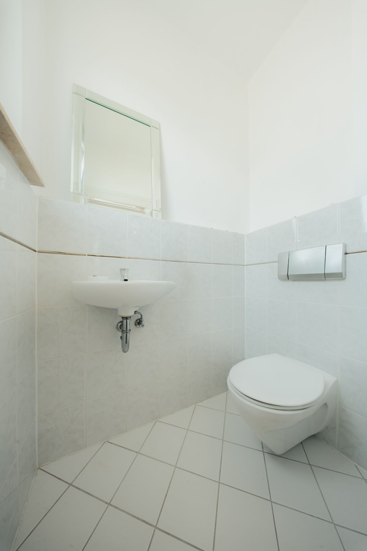 Kleines, weiß gefliestes Bad mit Wand-WC, Waschbecken und Spiegel.