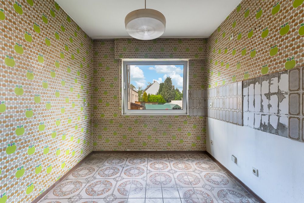 Leerer Raum mit gemusterter Tapete, Fenster und Fliesenboden, teilweise abgeblätterte Wandfliesen.