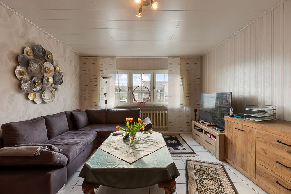 Wohnzimmer mit braunem Sofa, Holztisch mit Blumen, Fernseher und großem Fenster.