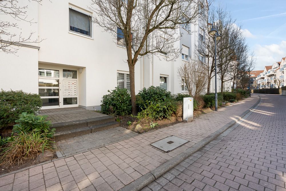 Eingang eines Mehrfamilienhauses mit Gehweg, Bäumen und Sträuchern davor.
