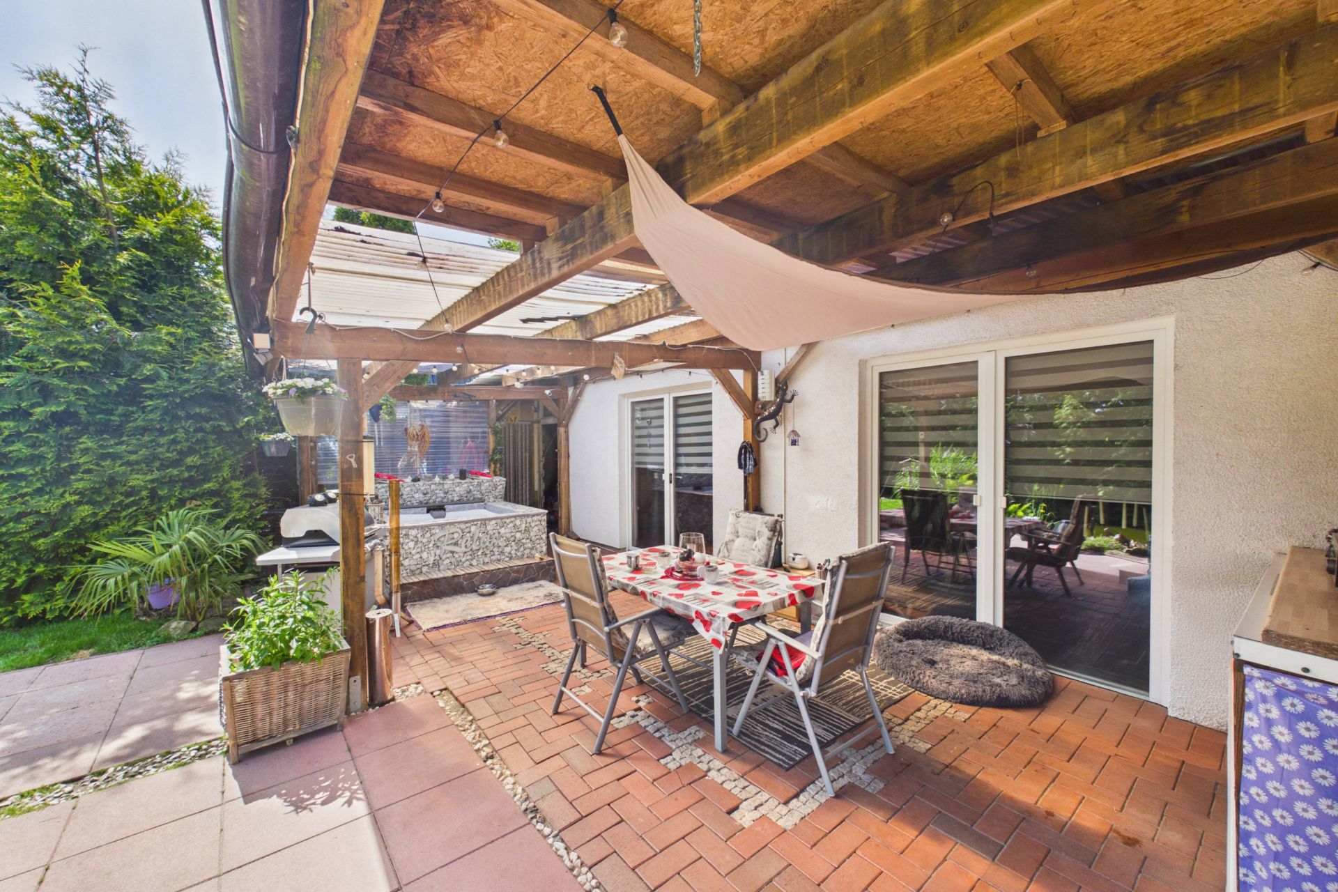 Überdachte Terrasse mit Esstisch, Stühlen und Zugang zu einem Garten, daneben eine Sitzecke.