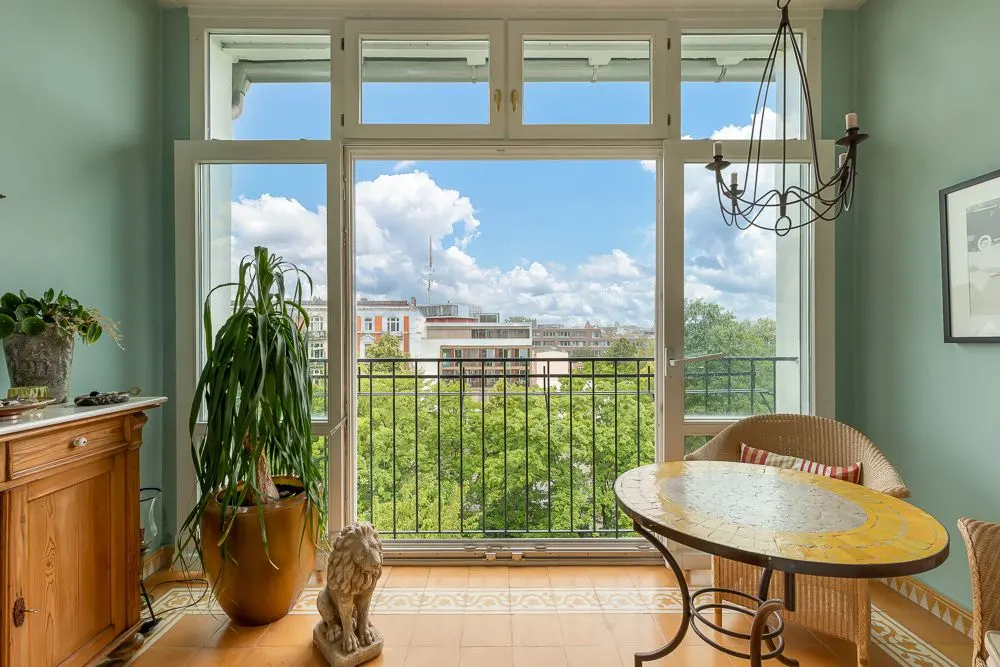 Heller Raum mit großem Fenster, Balkonblick ins Grüne, gelber Tisch, Rattanstühle und Pflanzen.