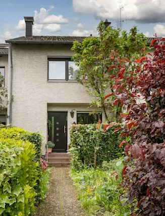 Zweistöckiges Wohnhaus mit Vorgarten, umgeben von verschiedenen Sträuchern und Pflanzen.