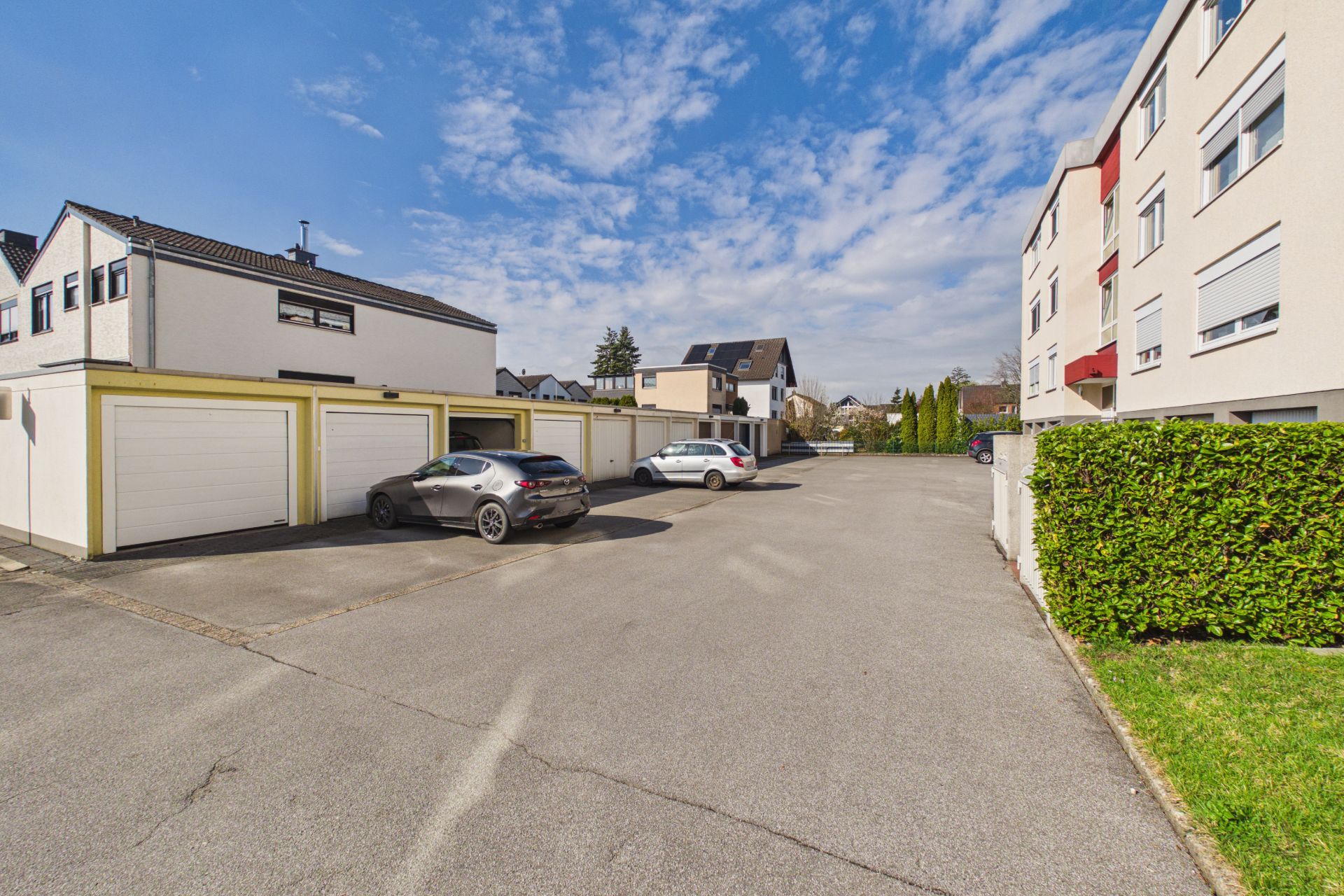 Asphaltierter Parkplatz mit zwei geparkten Autos vor Garagen und Wohngebäuden bei Tageslicht.