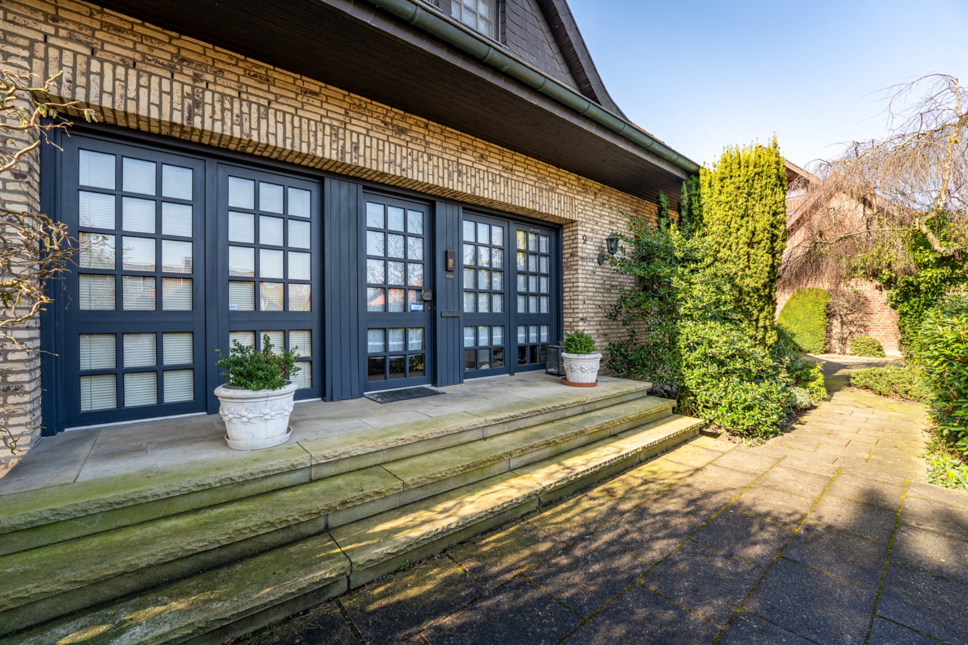 Eingang eines Wohnhauses mit breiten Treppen, großen Fenstertüren und zwei Blumenkübeln.