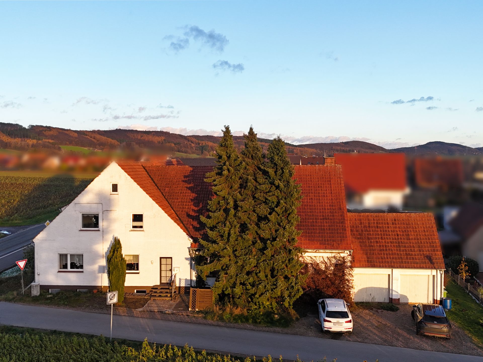 Weißes Wohnhaus mit rotem Dach, zwei Bäumen und parkenden Autos vorne.