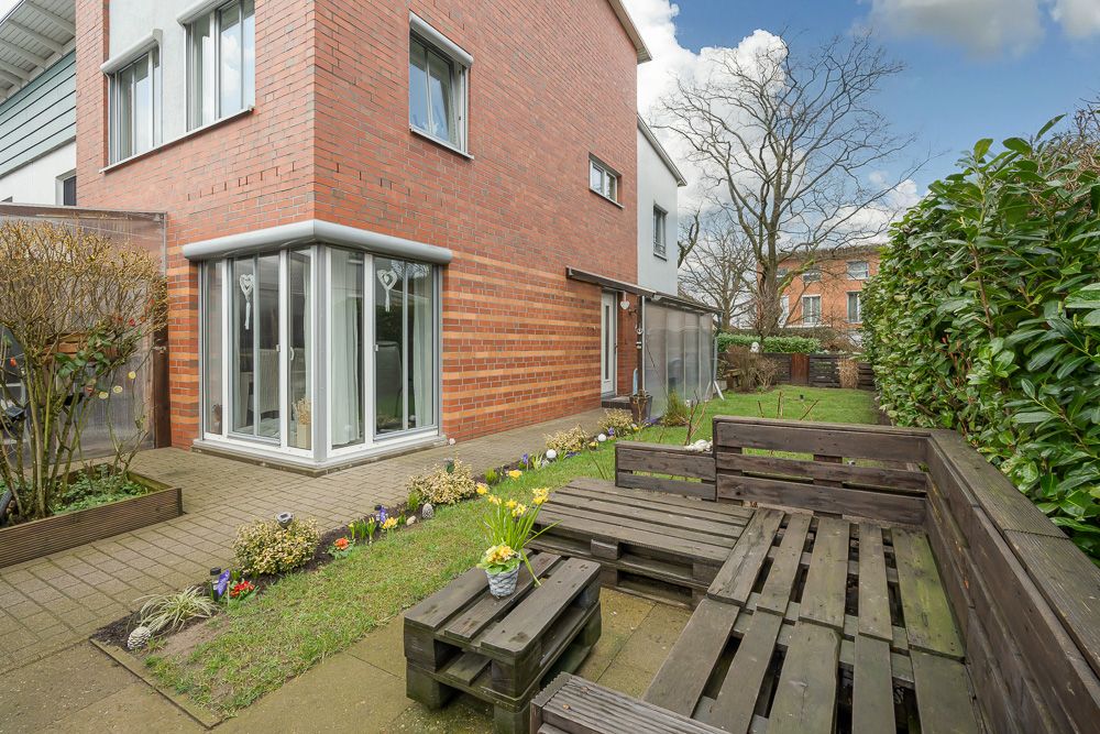 Moderne Terrasse mit Holzsitzgruppe und kleinem Garten neben rotem Backsteinhaus.