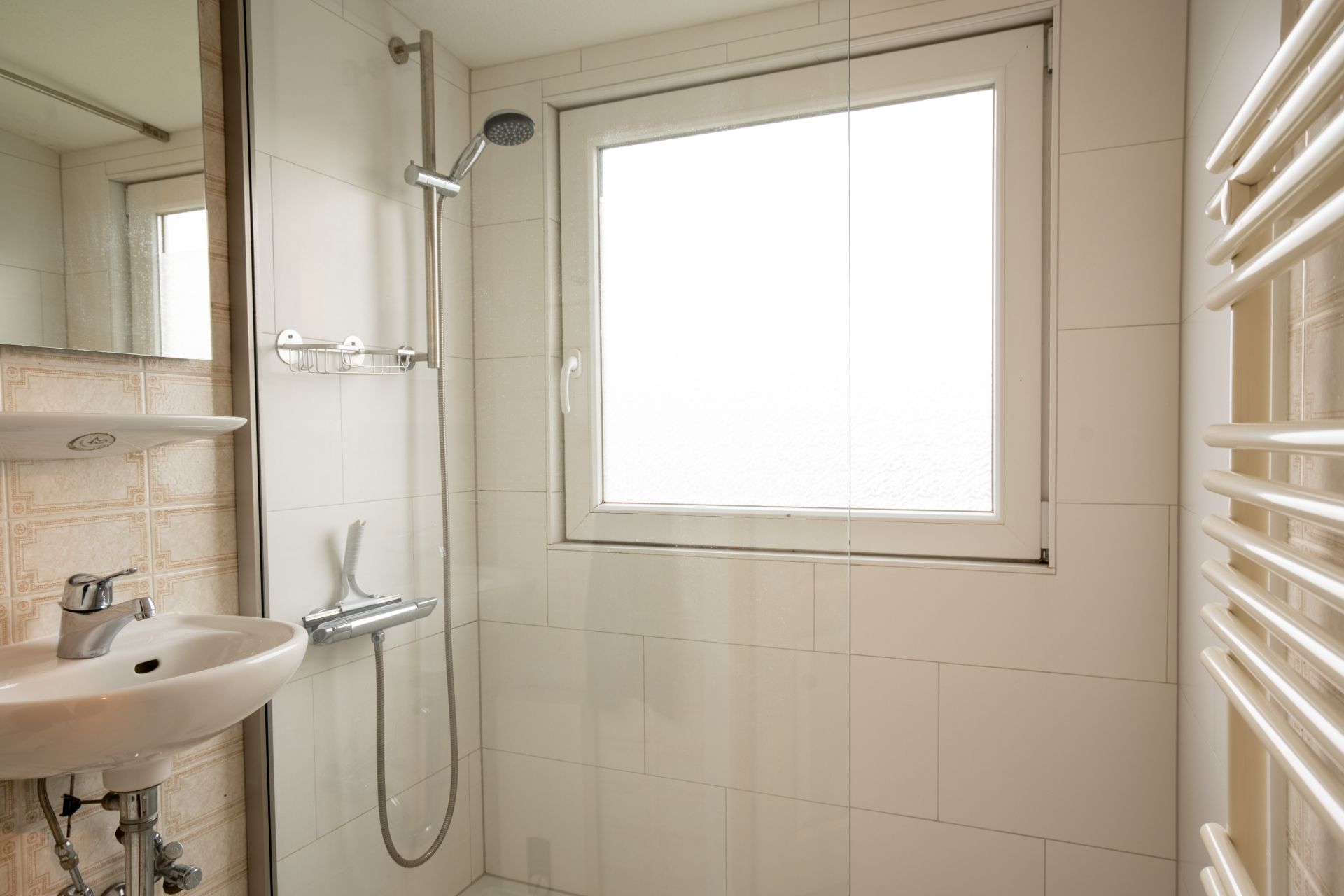 Helles Badezimmer mit Dusche, Fenster, Waschbecken und Handtuchheizkörper.
