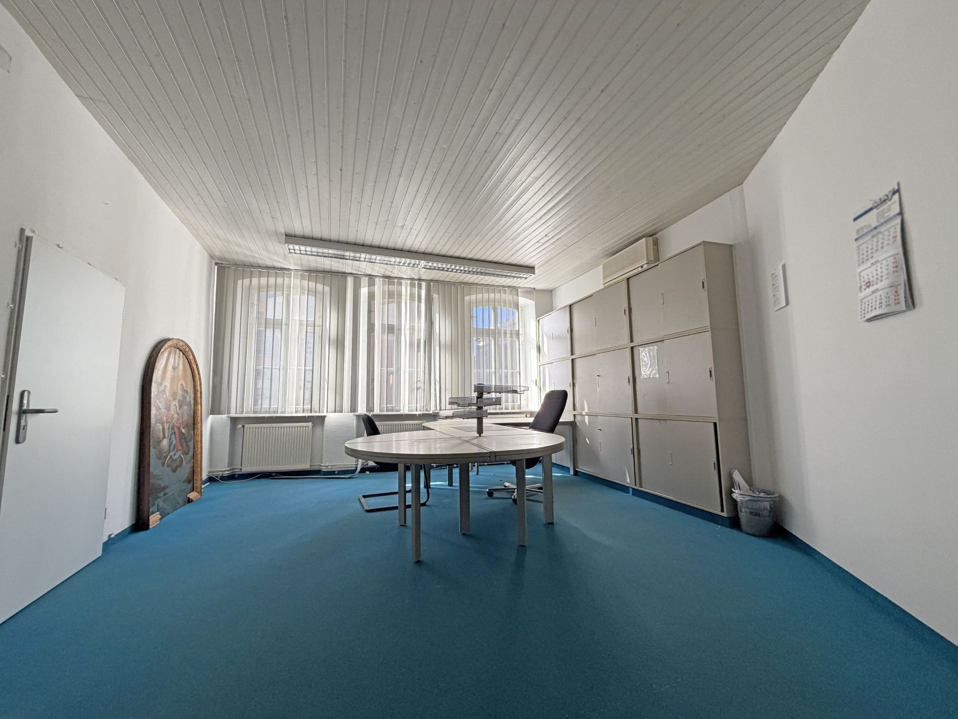 Helles Zimmer mit blauem Teppich, rundem Tisch, Stühlen und Aktenschränken an der Wand.
