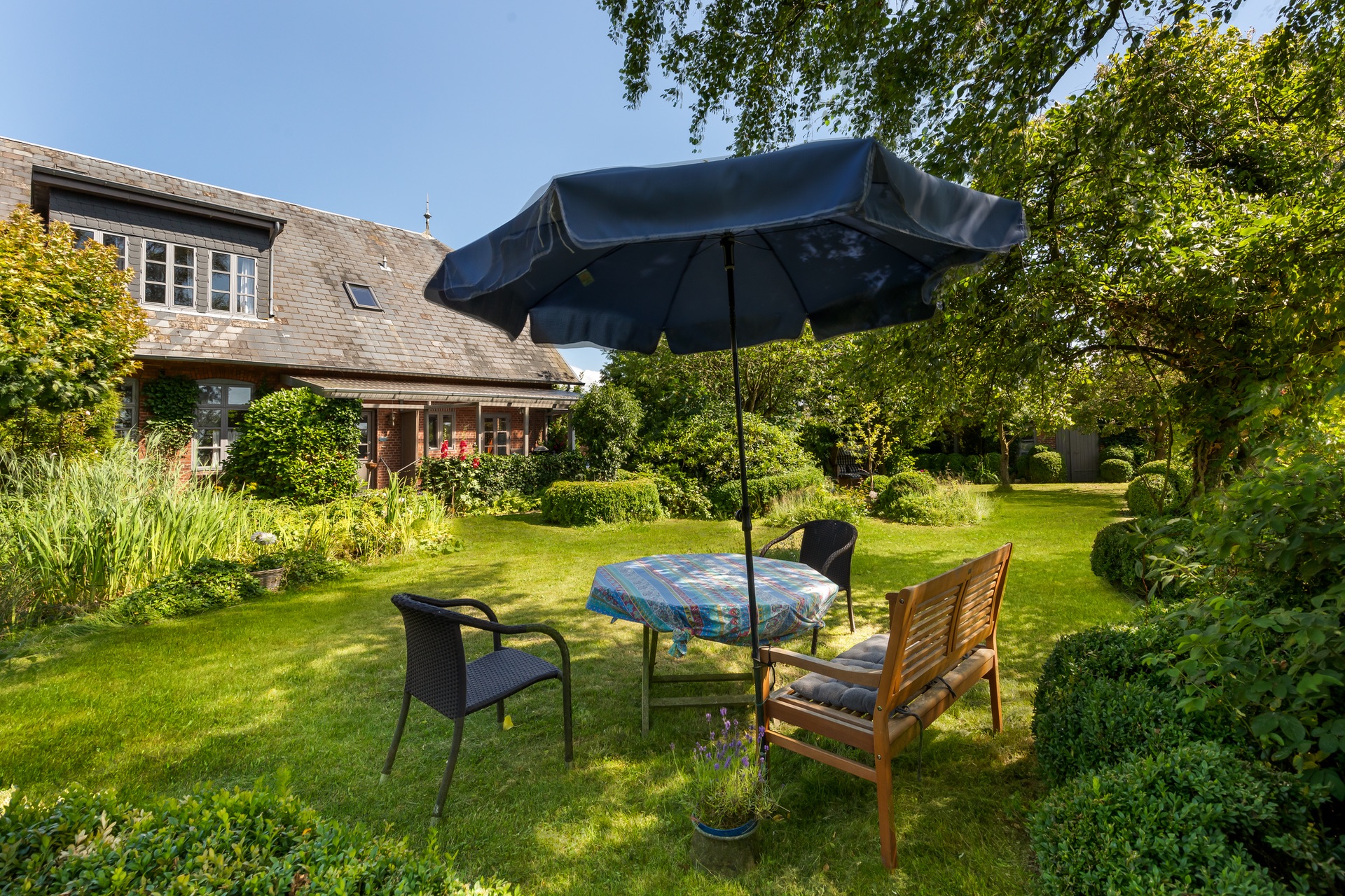 Garten mit Tisch, Stühlen und Sonnenschirm vor einem Wohnhaus an einem sonnigen Tag.