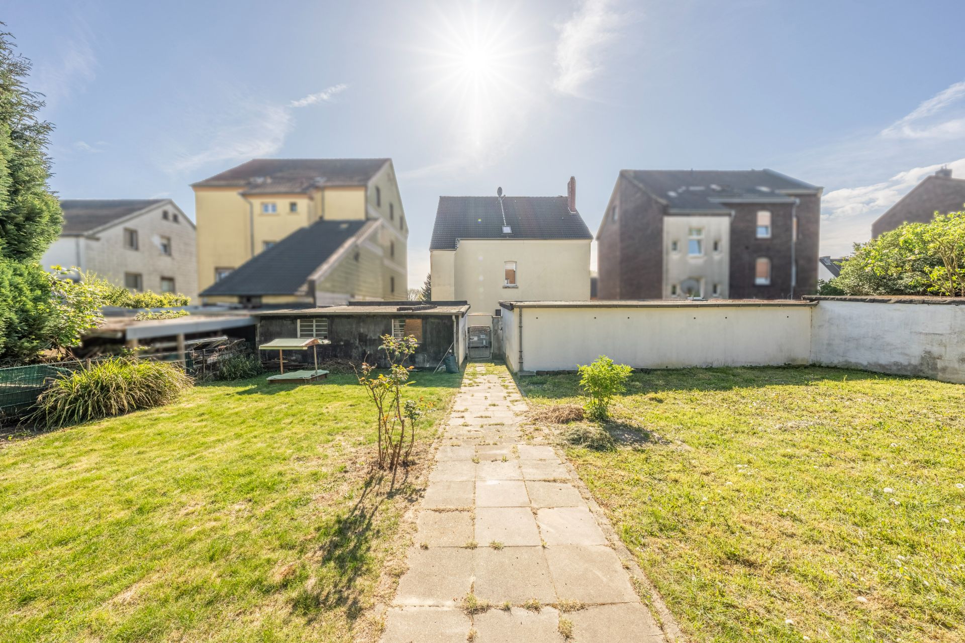 Gepflegter Garten mit Rasenfläche, Weg aus Steinplatten und angrenzenden Wohnhäusern bei Sonnenschein.