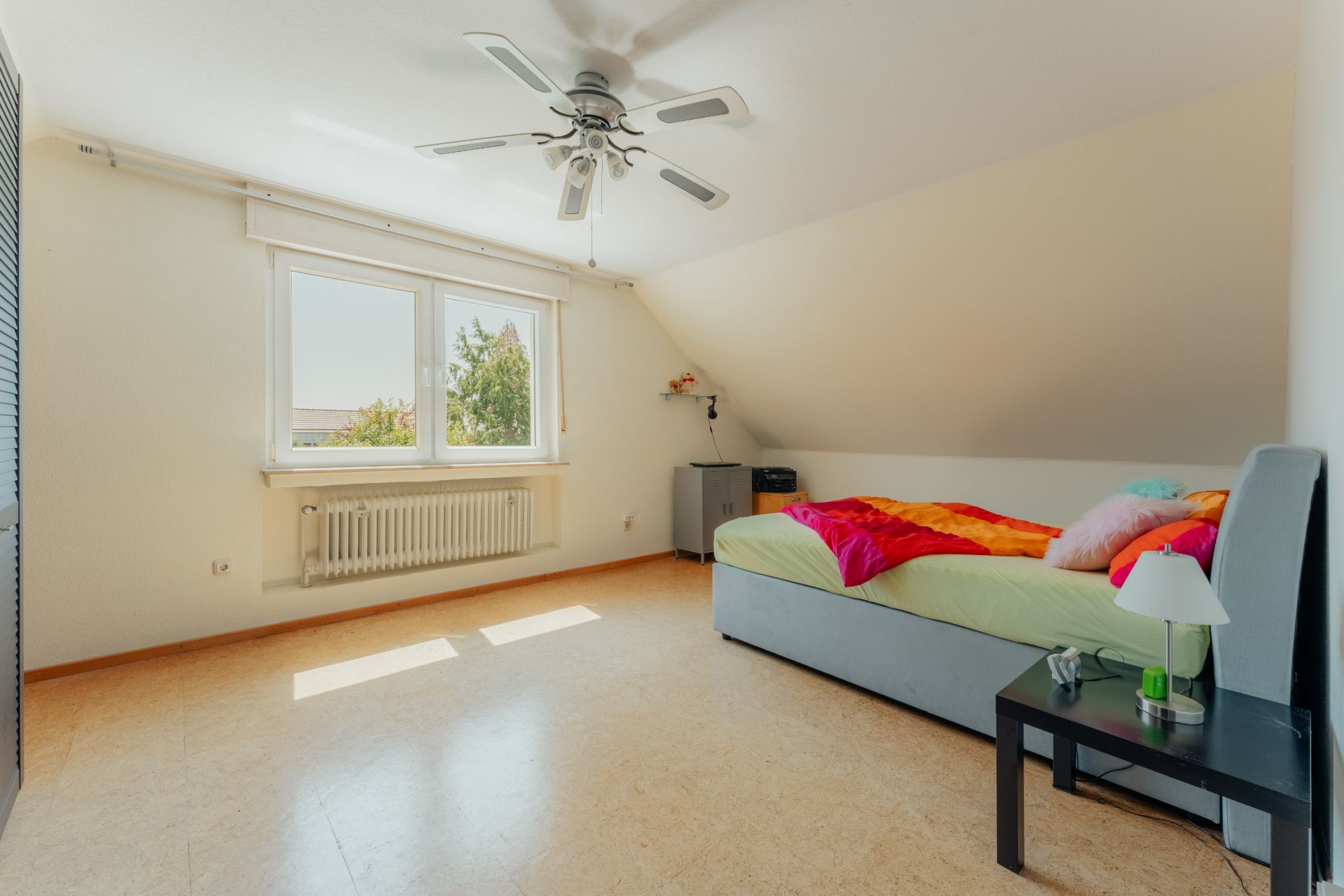 Helles Schlafzimmer mit Einzelbett, Nachttisch, Deckenventilator und großem Fenster.
