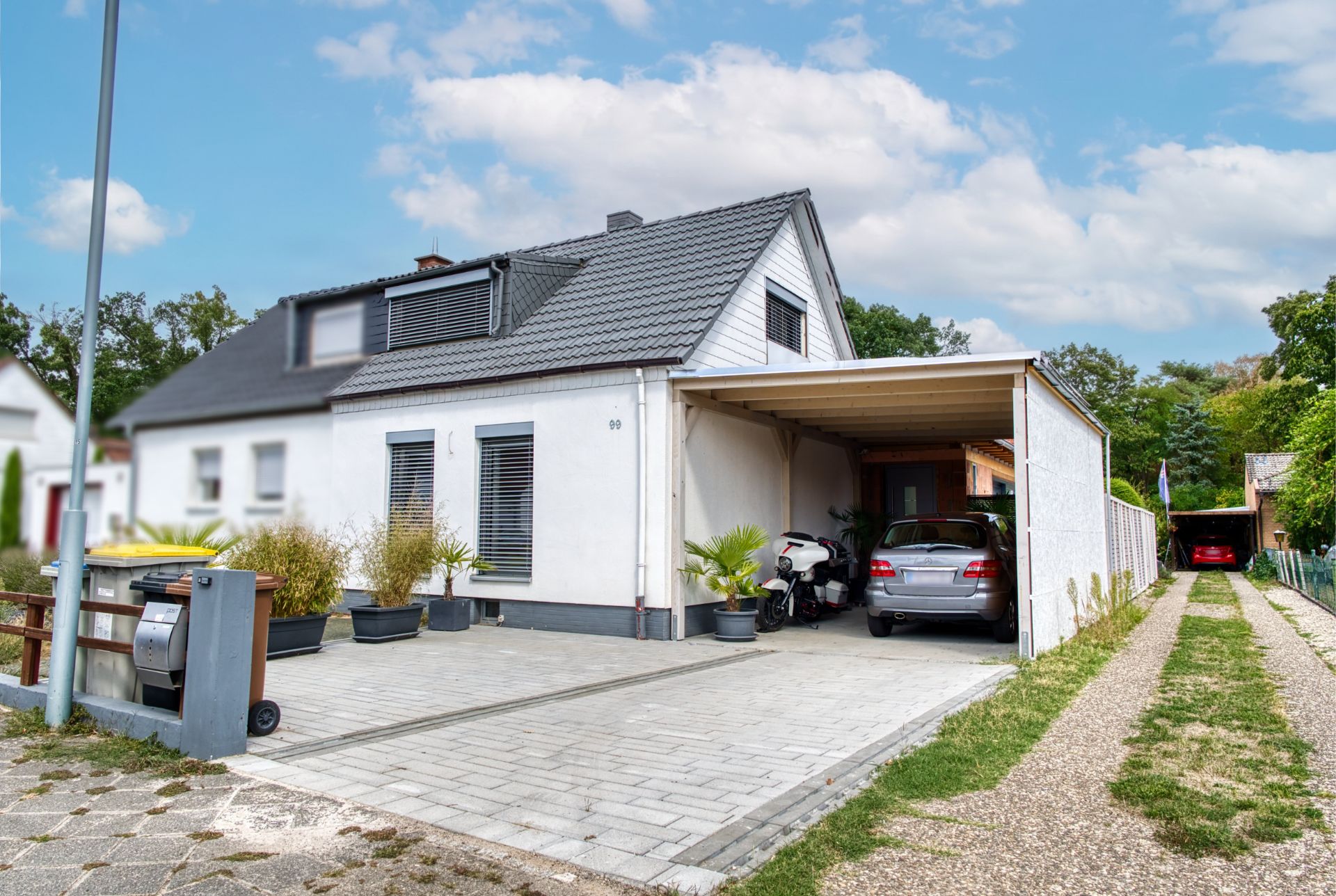 Modernes weißes Wohnhaus mit Carport, Auto und Roller auf gepflasterter Einfahrt.