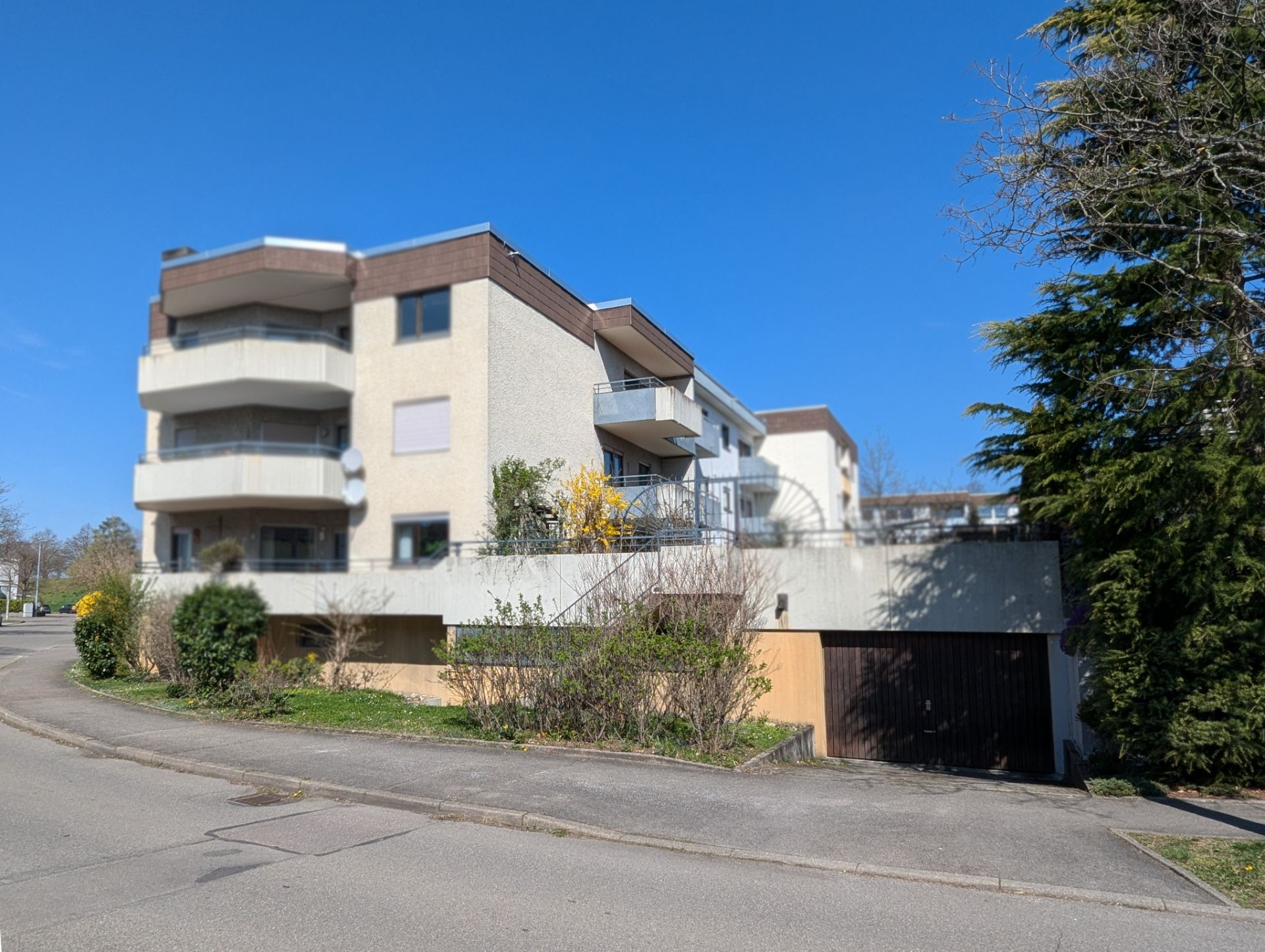 Mehrfamilienhaus mit Balkonen und Garage, umgeben von Büschen, an einer ruhigen Straße.