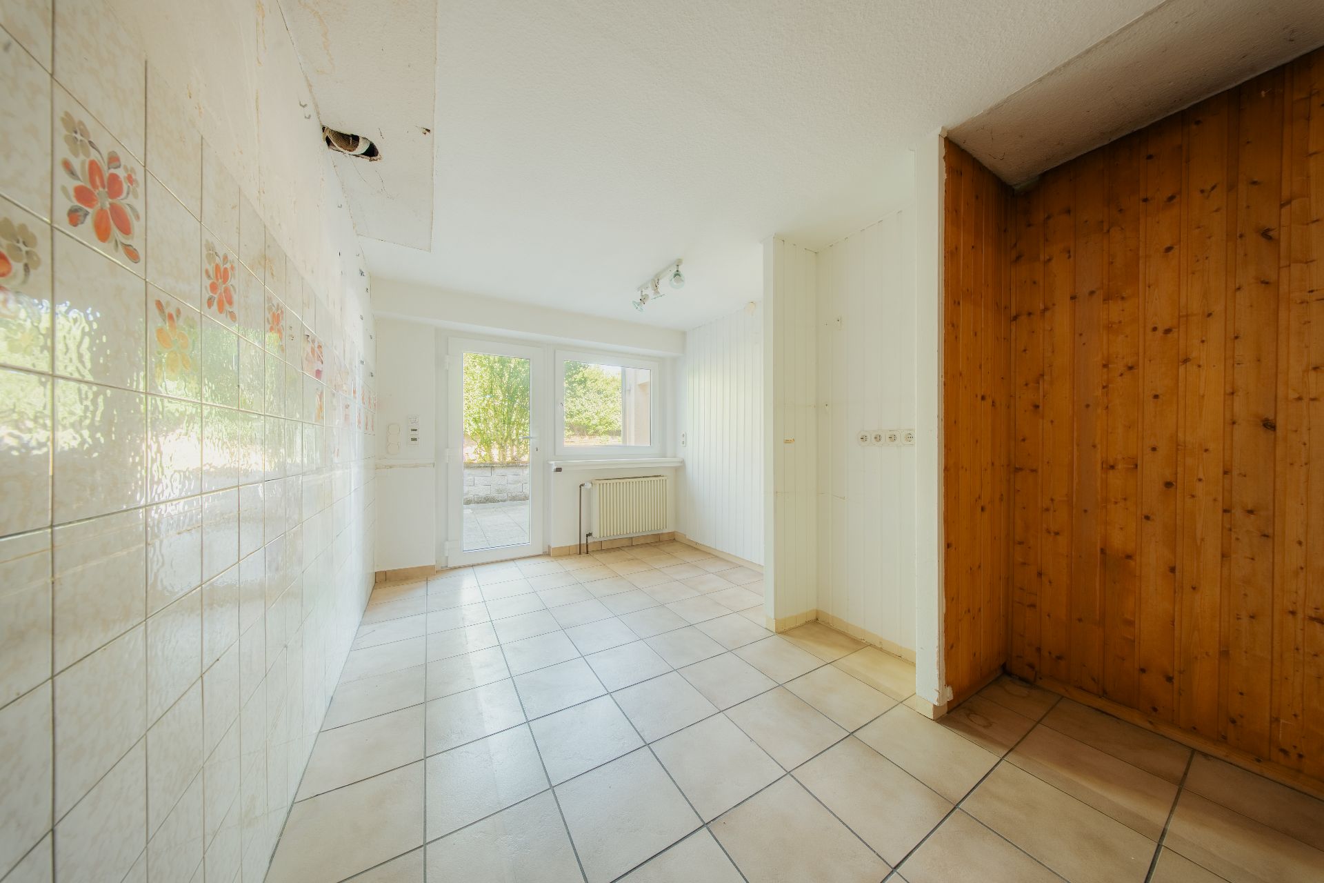 Leerer, heller Raum mit Fliesenboden, Holzwand und Fenster mit Blick ins Grüne.
