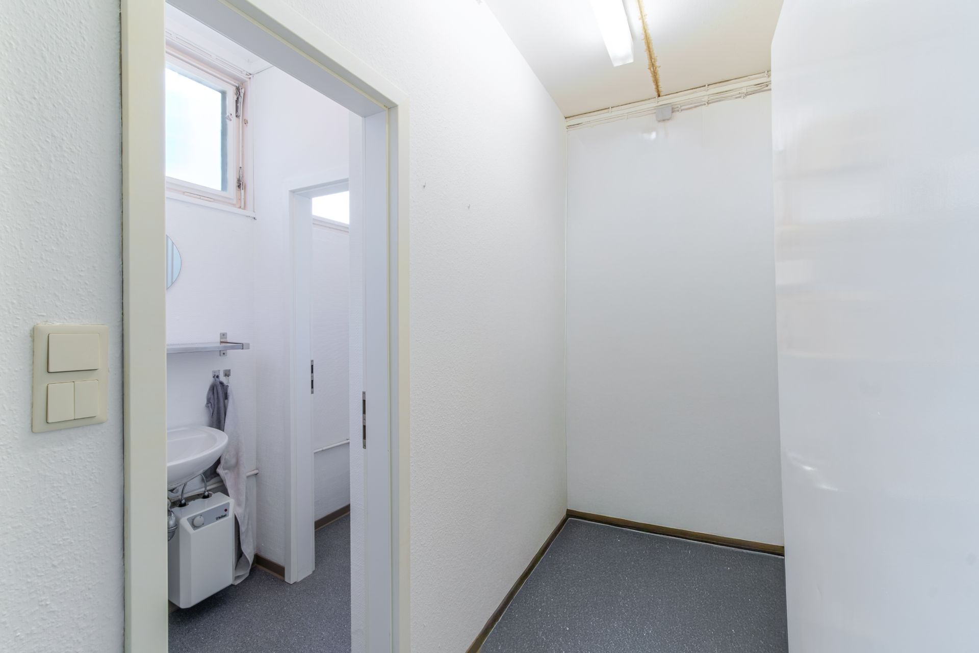 Flur mit grauem Boden, rechts eine weiße Wand, links Zugang zu einem kleinen Bad mit Waschbecken.