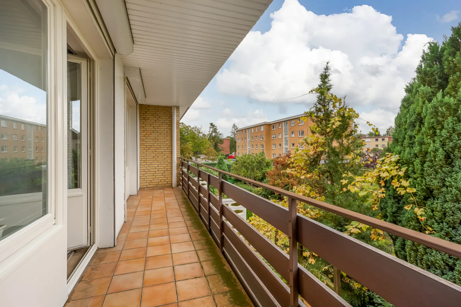 Langer Balkon mit braunen Fliesen, Ausblick auf Bäume und benachbarte Wohngebäude.