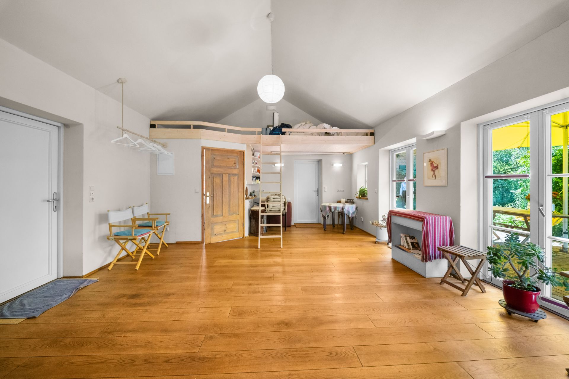 Helles Wohnzimmer mit Holzfußboden, Hochbett, großen Fenstern und Pflanzen.
