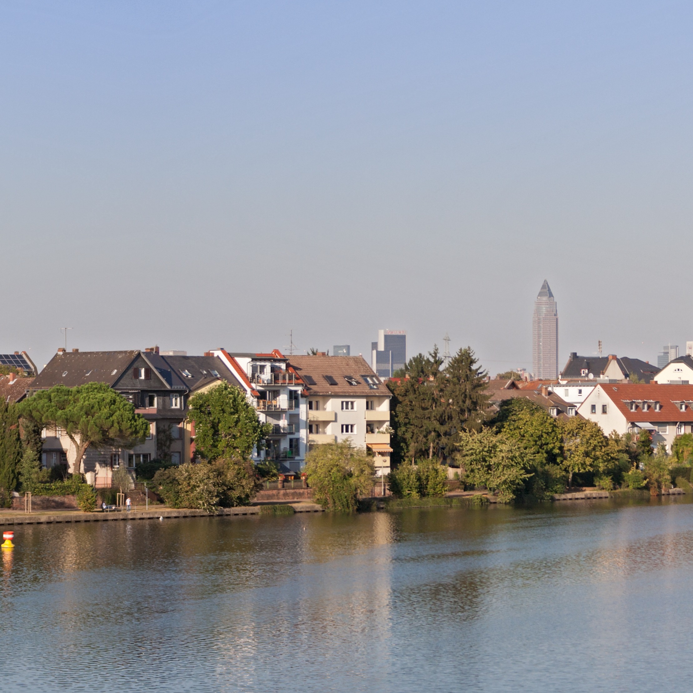 Mehrfamilienhäuser mit Balkonen am Flussufer, Bäume im Vordergrund, Stadtgebäude im Hintergrund.