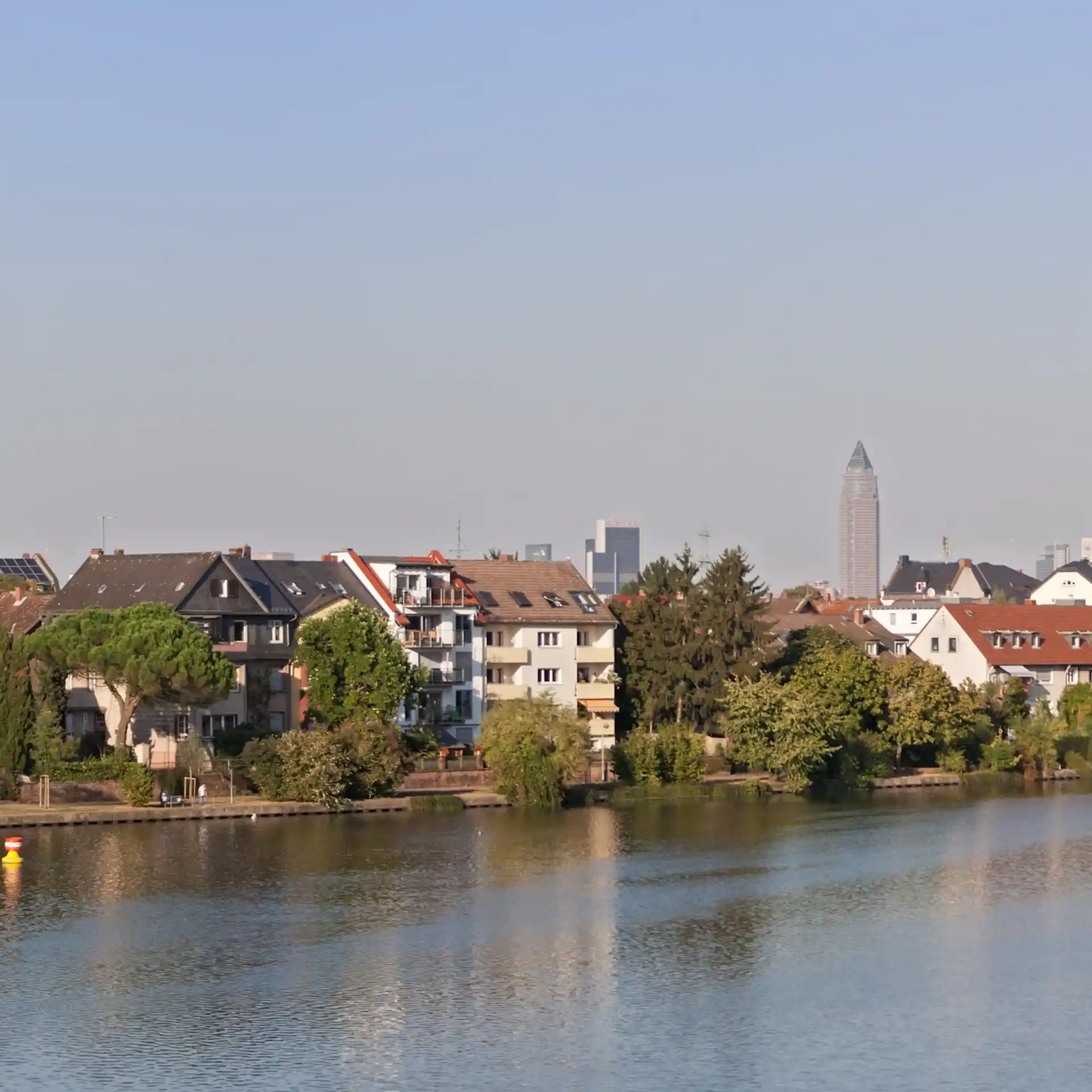 Mehrfamilienhäuser mit Balkonen am Flussufer, Bäume im Vordergrund, Stadtgebäude im Hintergrund.