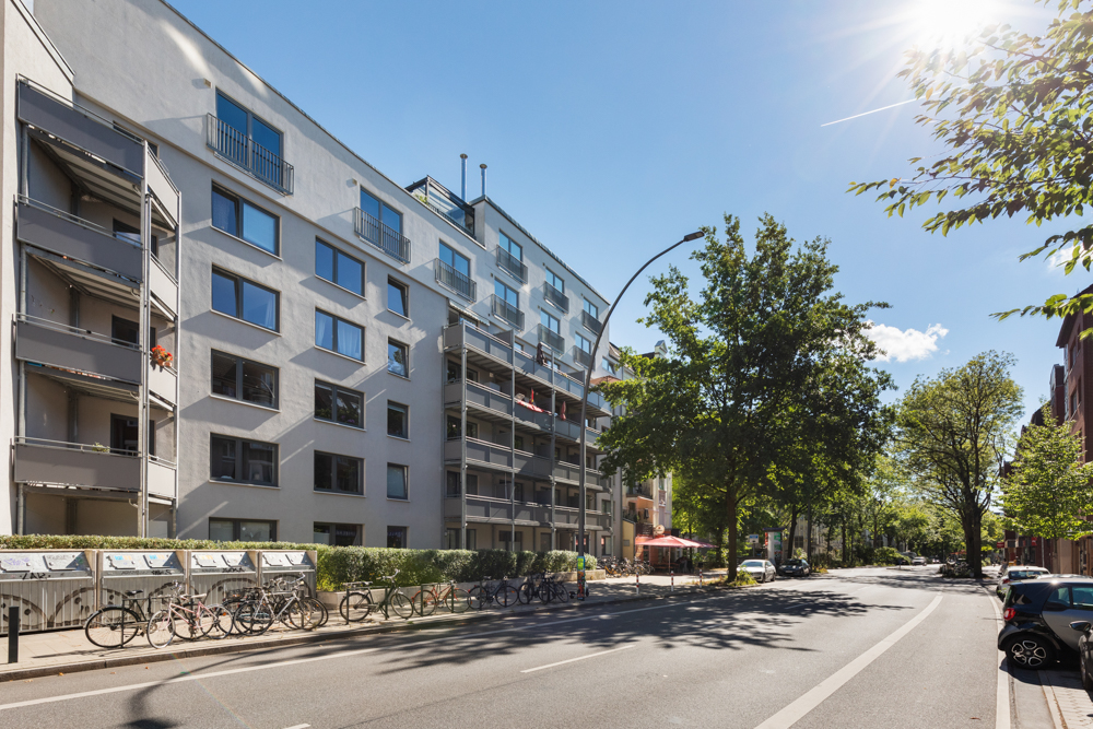 Mehrstöckiges Wohngebäude mit Balkonen, Fahrrädern vor dem Haus und Bäumen an einer sonnigen Straße.