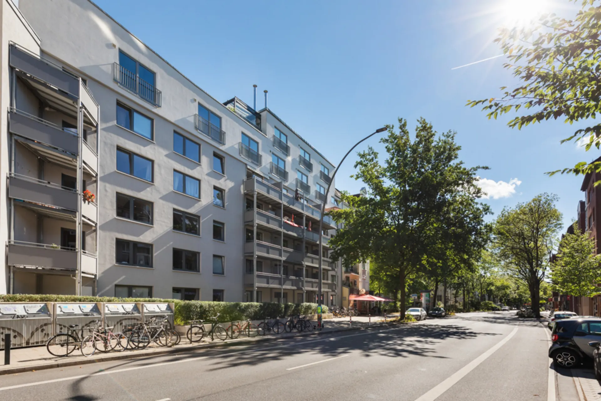 Mehrstöckiges Wohngebäude mit Balkonen, Fahrrädern vor dem Haus und Bäumen an einer sonnigen Straße.