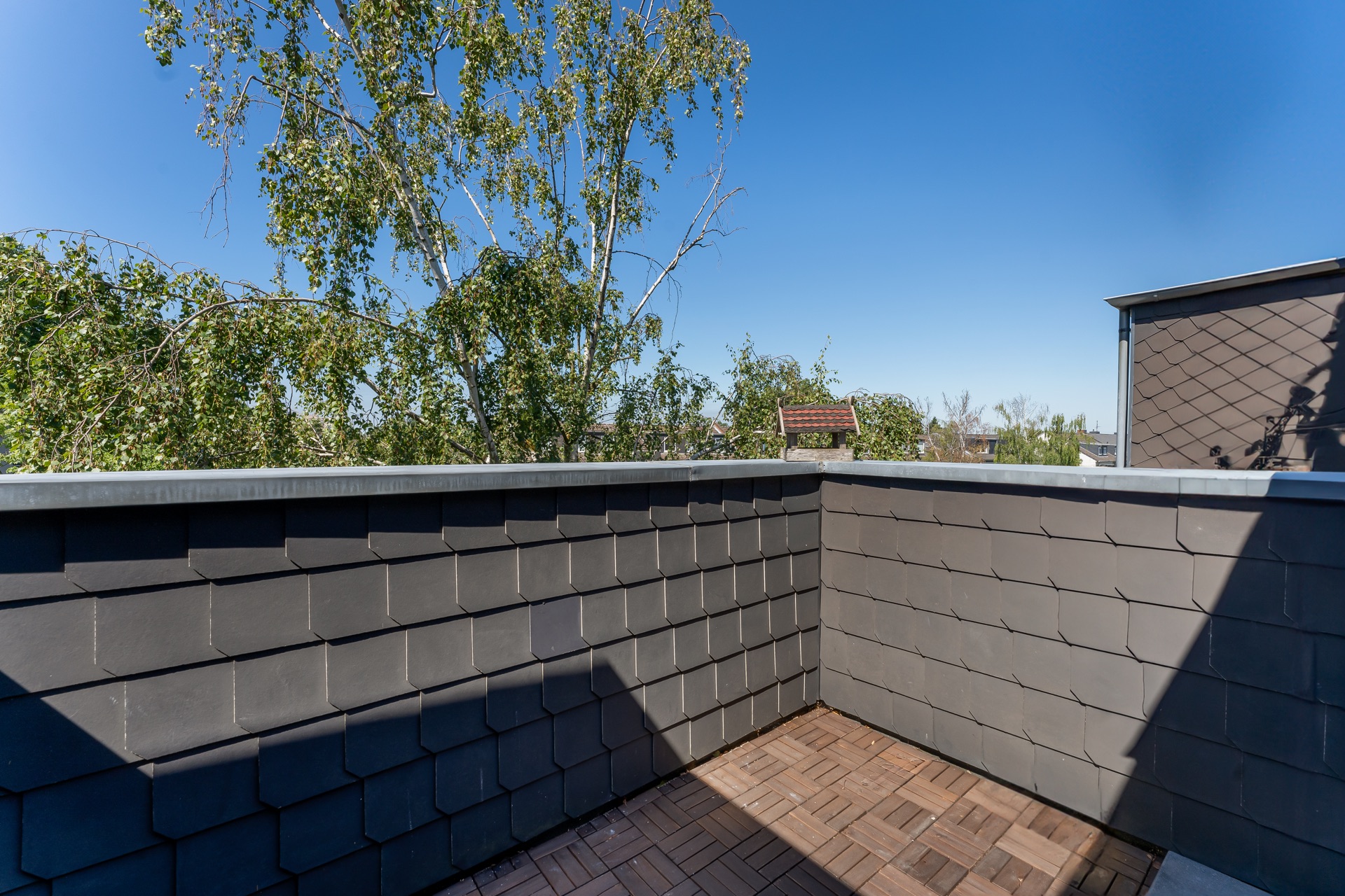 Kleine Dachterrasse mit Holzboden und Blick auf Bäume bei klarem Himmel.