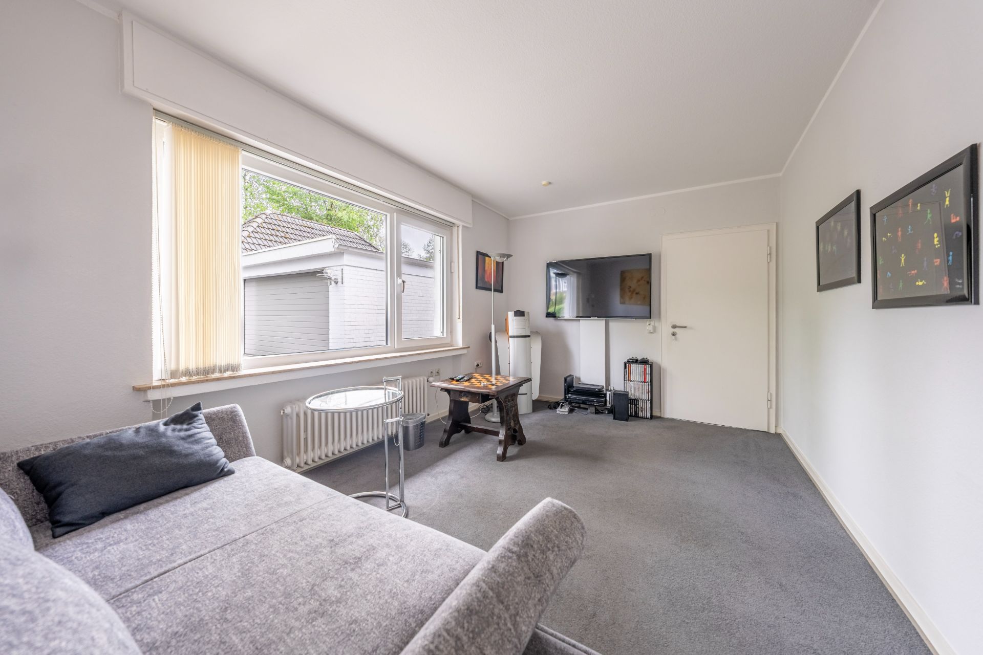 Helles Wohnzimmer mit grauem Sofa, Glastisch, großem Fenster und TV an der Wand.