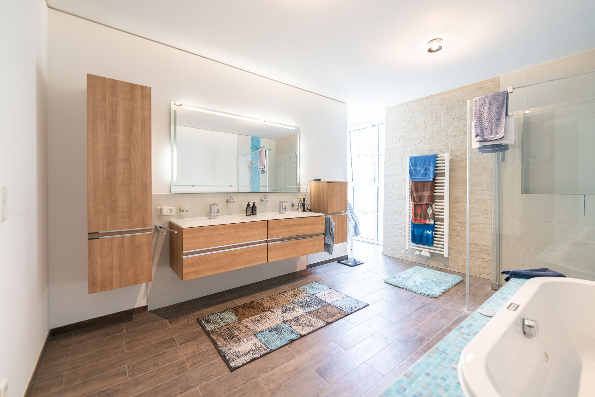 Modernes Badezimmer mit Holzschränken, großem Spiegel, Badewanne und Dusche auf Fliesenboden.