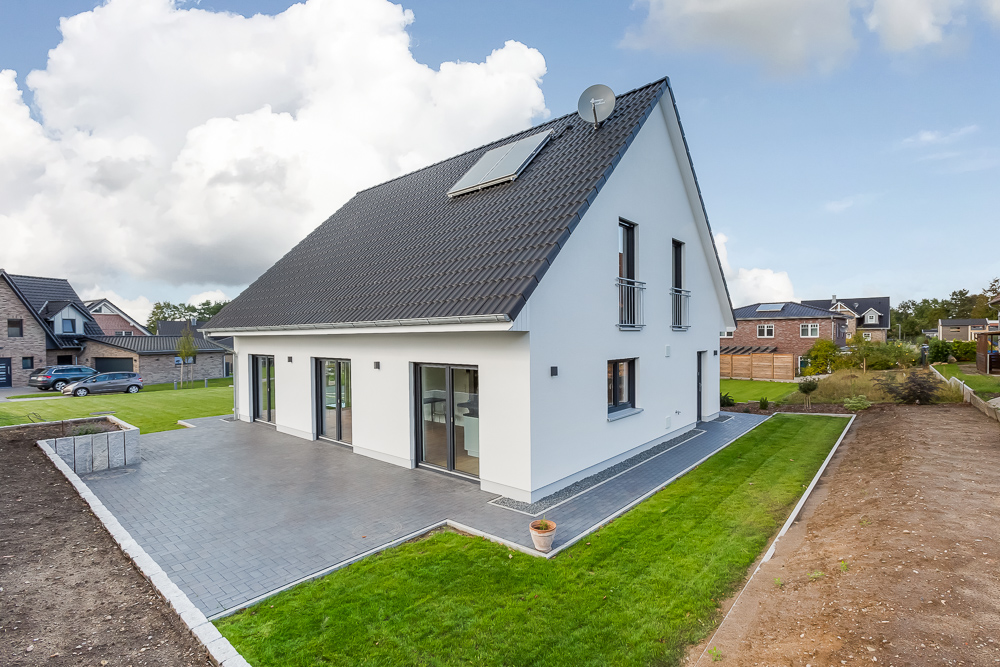 Modernes weißes Einfamilienhaus mit Garten und gepflasterter Terrasse bei Tageslicht.