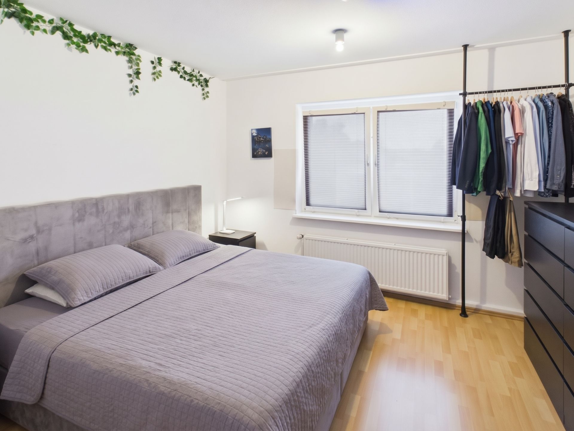 Schlafzimmer mit Doppelbett, Fenster, Kleiderstange und Kommode auf Laminatboden.