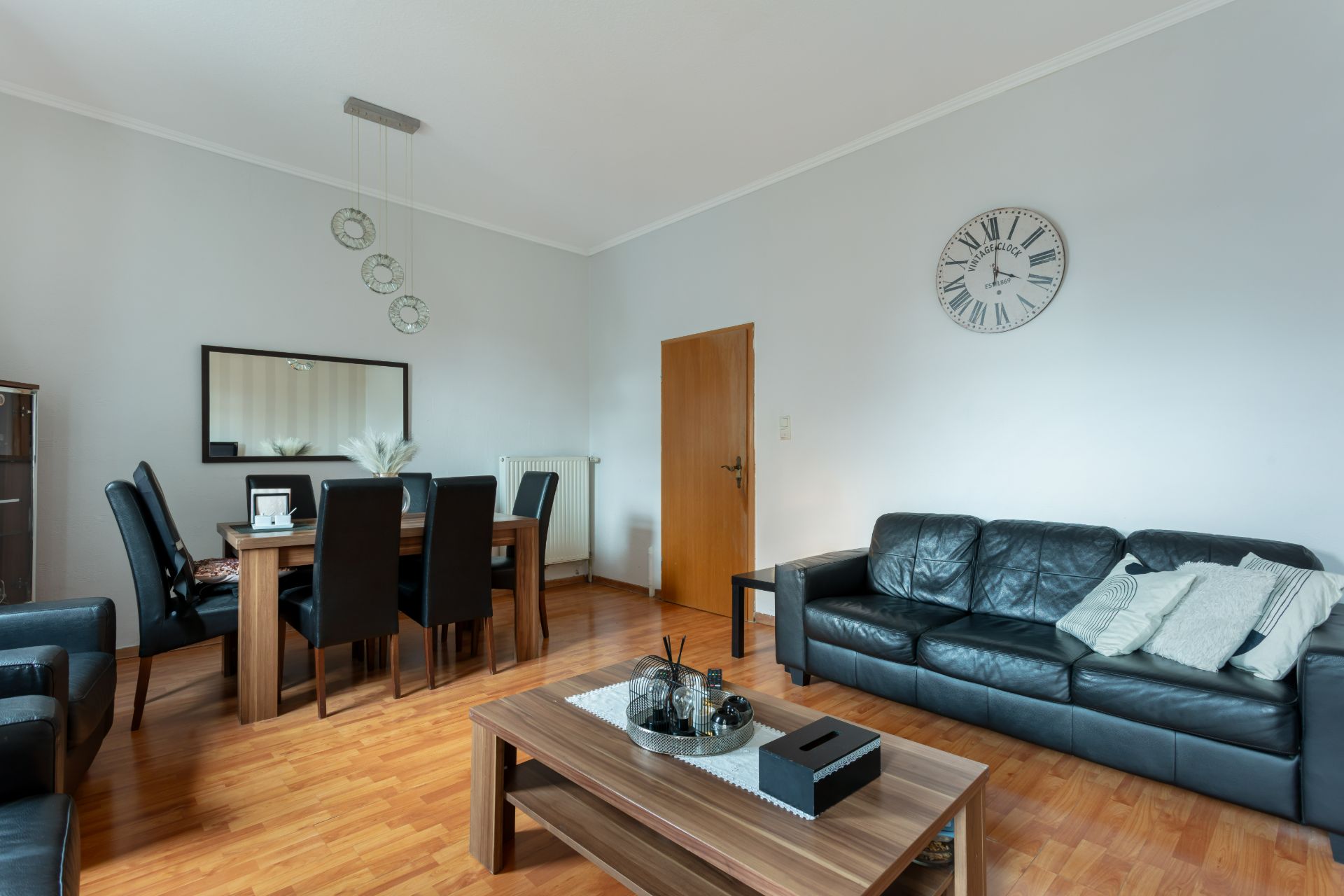 Wohnzimmer mit schwarzem Ledersofa, Esstisch, sechs Stühlen, Couchtisch und großer Wanduhr.
