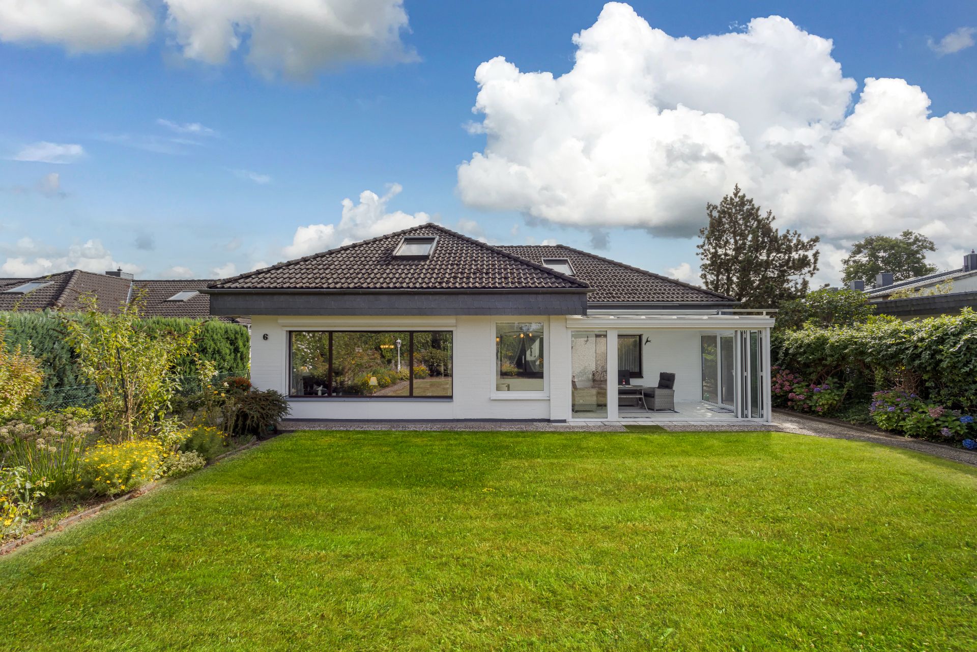 Einfamilienhaus mit großem Garten, Terrasse und Glasfront, umgeben von Hecken und Blumen.
