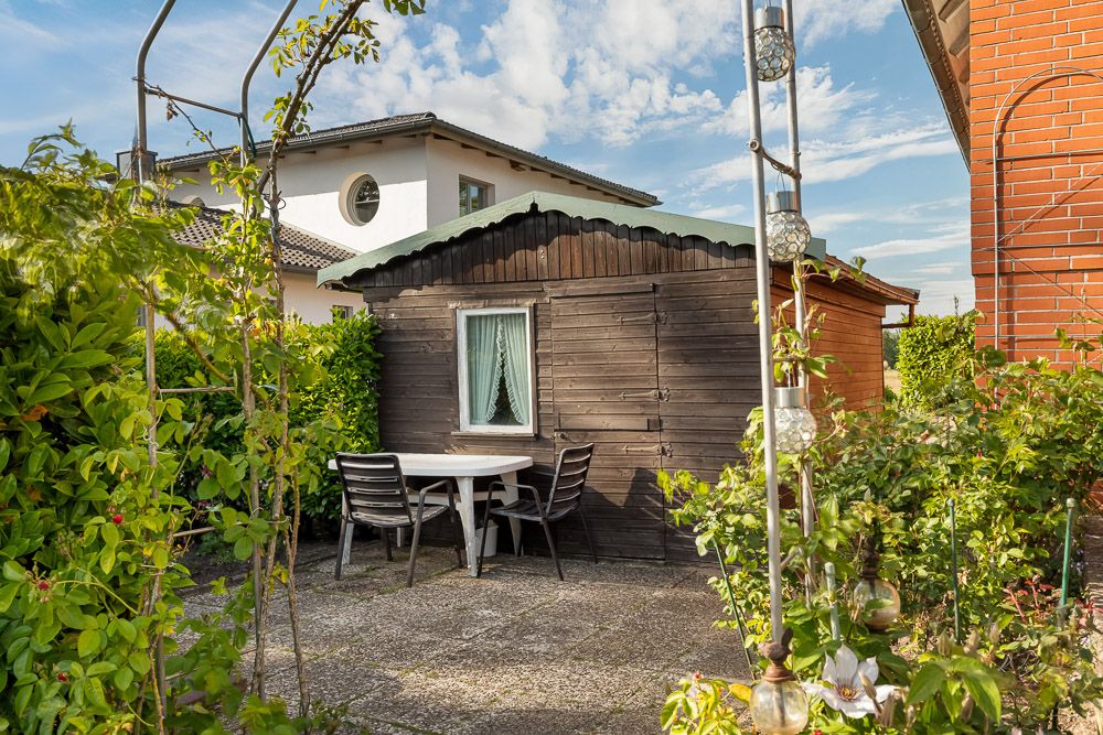 Garten mit kleinem Holzhaus, rundem Tisch und drei Stühlen auf einer Terrasse.